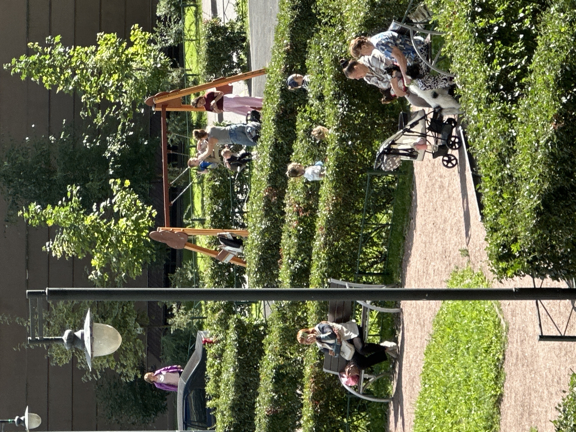 The image depicts an outdoor scene in the city of Murino, Russia, on a sunny day. In the foreground, a person is seated on a park bench, holding a phone to their ear. Next to them is a light-colored bag. To the right, two women are seated on a bench, one wearing a patterned shirt and the other a blue floral shirt. A dog with black and white markings is standing near them. A baby stroller is parked nearby.
In the midground, a playground with a swing set is visible. Several children and adults are present, with one child on a swing and others interacting around the playground. A black car is parked to the left, behind a neatly trimmed hedge.
The background features tall trees and a building with a dark facade. A lamppost stands prominently in the left-center of the image. The environment is characterized by lush green grass, well-maintained hedges, and a gravel path. The overall impression is of a public park or residential courtyard.