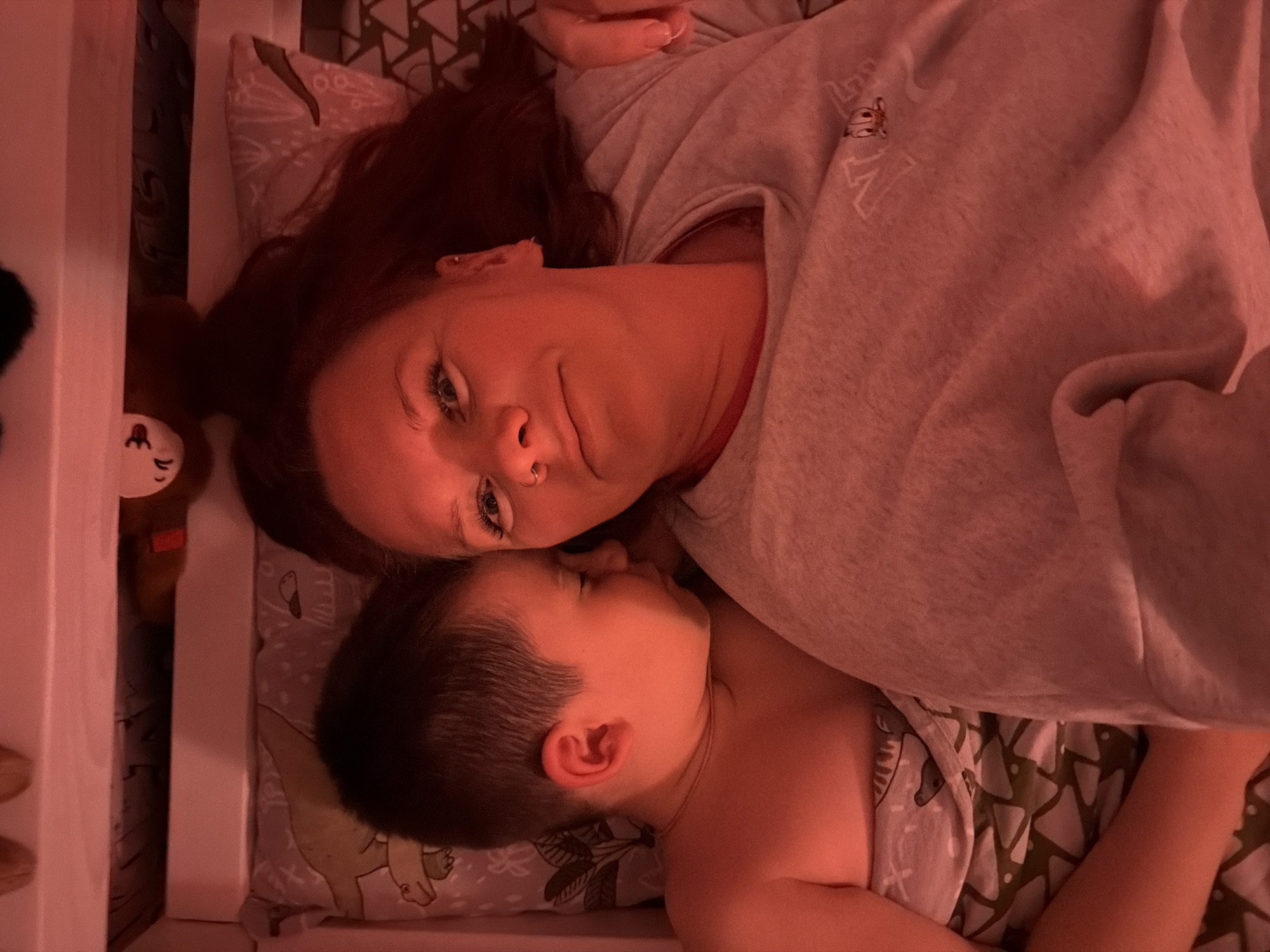 The image depicts a tender and intimate moment between a woman and a young child, presumably a mother and her son, lying together in a dimly lit, warm environment, likely a bedroom in Chaikovsky, Russia. The scene is illuminated by a predominant reddish-orange ambient light, suggesting either evening or nighttime.
The woman, positioned above and to the left of the child, has auburn or dark red hair and is looking towards the camera with a neutral to slightly contemplative expression. She has visible blue eyes, long eyelashes, and a nose piercing. She is wearing a light-colored (possibly grey or faded pink under the lighting) sweatshirt, on which a small embroidered design is visible on the chest. This design includes what appears to be partial letters, possibly 'N' and 'E', alongside a small character resembling a frog or turtle.
The young child, with dark hair, is nestled closely to the woman, sleeping peacefully with eyes closed. The child appears shirtless, revealing their shoulder and ear. Both are lying on pillows covered with patterned cases; one visible pattern includes a cartoon-like dinosaur, and another features abstract shapes. A patterned blanket or sheet is also visible. On the far left of the frame, a white crib railing is visible, suggesting a co-sleeping arrangement or a child's bed. Partially obscured by this railing is a small brown stuffed animal with a light-colored face. The overall atmosphere conveys a sense of peacefulness, comfort, and maternal bond.