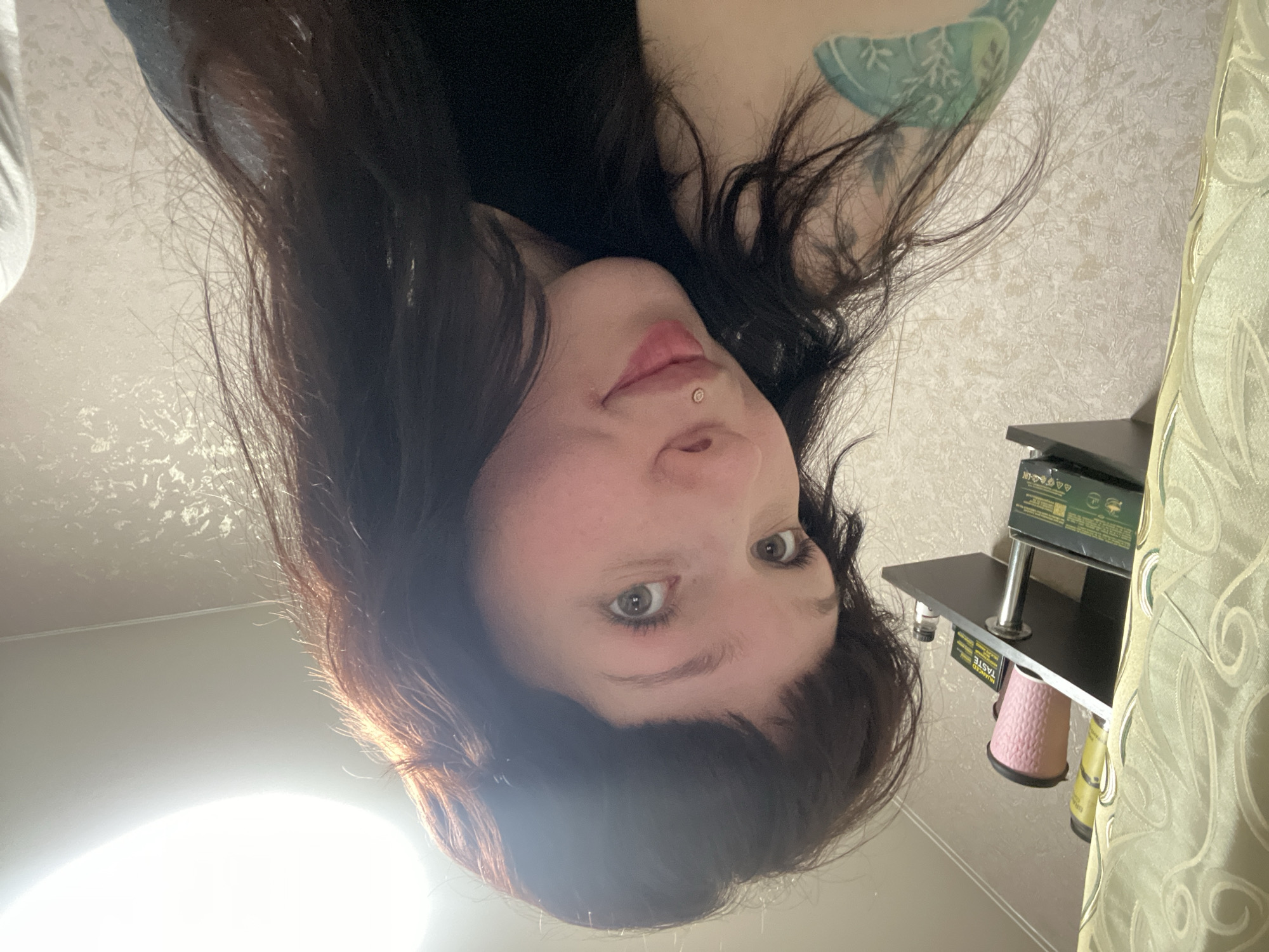 A female individual with fair skin, dark brown hair, and light-colored eyes is seen in the image, facing the camera at an upside-down angle. She has a piercing above her upper lip. Part of a tattoo is visible on her upper arm. The background shows a textured wall, a curtain, and a shelf with objects on it including what appears to be a pink cup. Although the image cannot be explicitly traced to the city of Chaikovsky, Russia without additional evidence, the setting appears to be indoors, possibly in a domestic environment, considering the items visible. No specific actions or interactions are discernible.