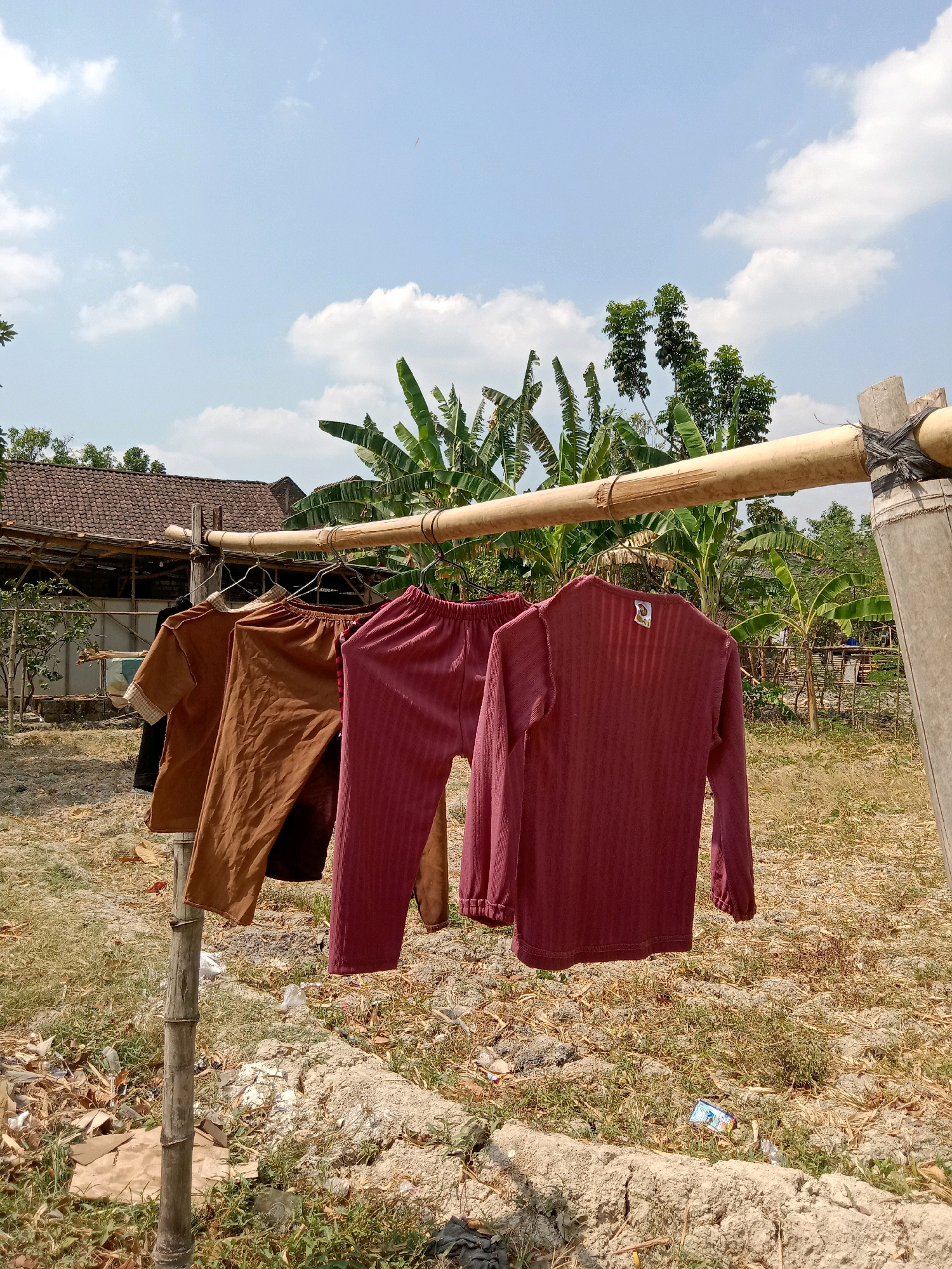 The image depicts laundry hanging on a bamboo clothesline outdoors. The scene appears to be in a rural area, possibly the city of Stiaji, Indonesia, given the presence of banana trees and a traditional tiled roof house in the background. The clothesline is supported by two bamboo poles. Several pieces of clothing, including two long-sleeved shirts and what appear to be pants or pajamas, are hung on the line. The ground is covered with dry grass, leaves, and some scattered debris. The sky is blue with scattered white clouds, indicating a sunny day. There are no people visible in the image. The overall impression is one of domestic life in a tropical or subtropical setting.