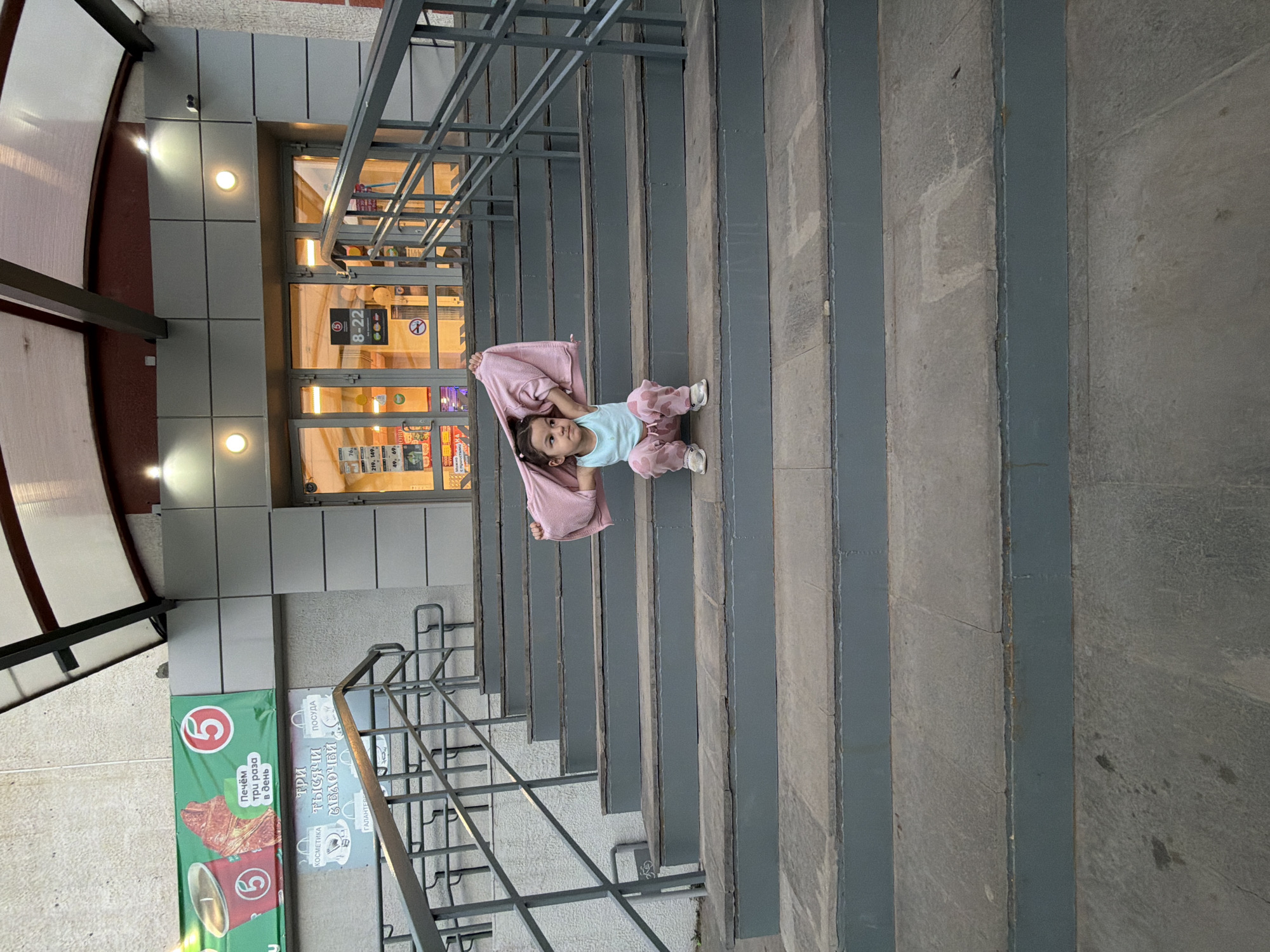 Here is a detailed description of the image:
**Scene Overview:**
The image shows a young child lying on a staircase outside what appears to be a commercial building. The perspective is slightly angled from above.
**People:**
* A child is lying on the stairs. The child wears a light blue top, pink patterned pants, and white shoes. The child holds a pink blanket overhead.
**Objects:**
* A long outdoor staircase with metal railings on either side.
* A pink blanket held by the child.
* Building with windows and doors
* Signage on the building related to a "5" retail business. Signs show food items and the phrase "печём три раза в день", which translates to "baked three times a day". There are also signs for cosmetics, dishes, and other products.
**Environment:**
* The scene is outdoors in a city-like environment.
* The lighting is bright, likely during daylight hours.
**Location Information:**
* Based on the signage, the location is Chaikovsky, Russia, as the "5" logo is known in that area. The signs are written in Russian.