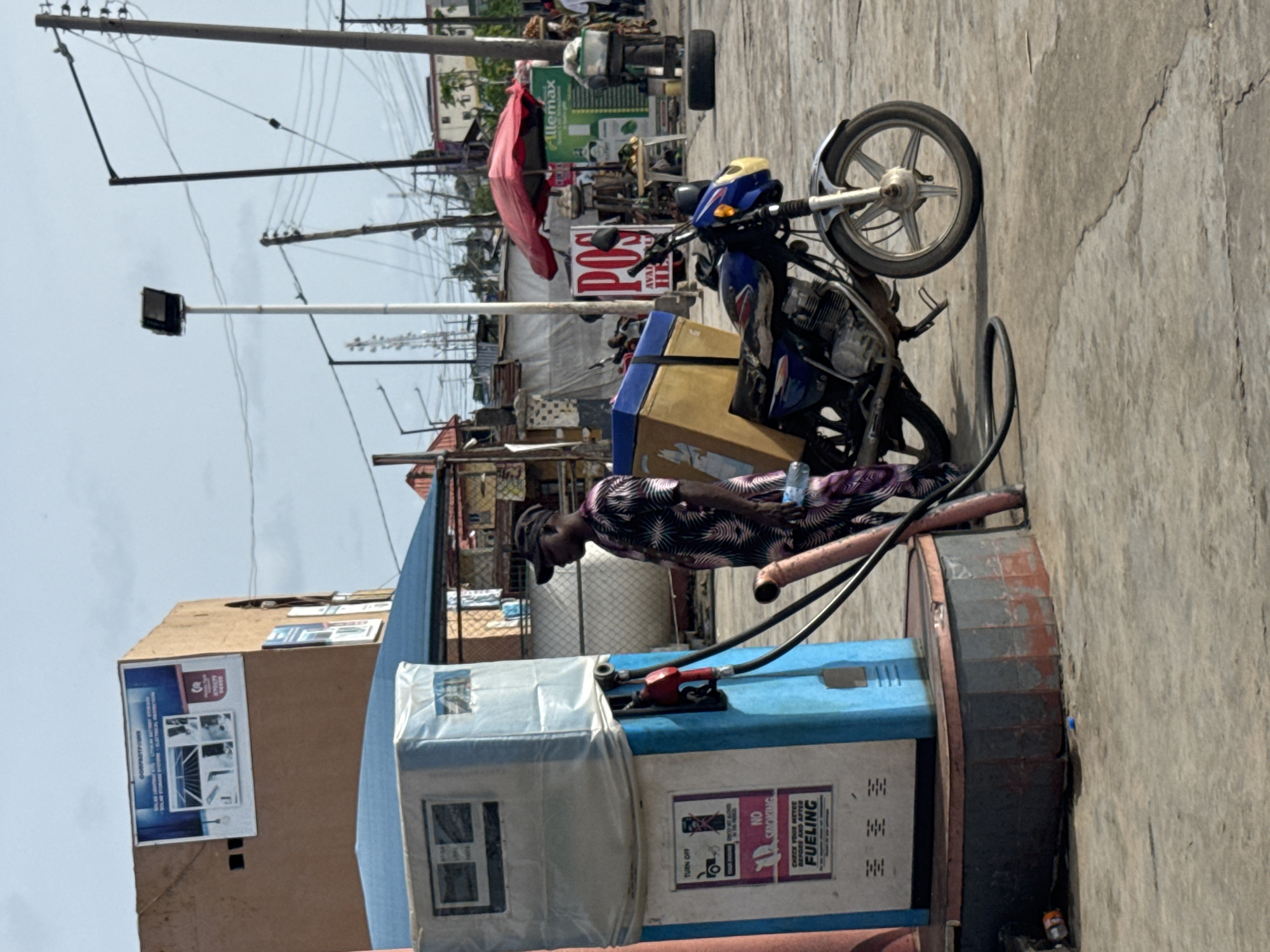 A vertical shot depicts a man, appearing to be in the act of fueling a motorcycle at what appears to be a roadside gas station in Badagry, Nigeria. The man, wearing a dark patterned shirt and a cap, is positioned next to a blue and white fuel dispenser that has multiple warning stickers. The dispenser is connected by a hose to a blue motorcycle, which has a yellow box attached to it. Behind the man and the motorcycle, there are structures and signs, including one that reads "POS AVAILABLE HERE". The ground is paved, and in the background, utility poles with electrical wires are visible, adding to the urban atmosphere.