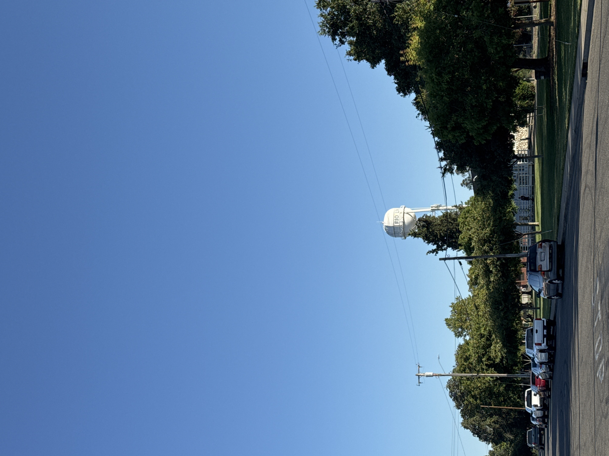 The image displays an outdoor scene, rotated 90 degrees clockwise, depicting a clear day in what appears to be a suburban or semi-rural area. The dominant feature is a large, white spherical water tower located towards the center-right of the frame, partially obscured by trees. The text "RIO L" is visible on the side of the water tower, consistent with the provided location context of Rio Linda.
A vast, unblemished blue sky occupies the upper two-thirds of the image, suggesting clear weather and ample daylight, likely midday or early afternoon given the brightness. Below the sky, a line of lush green trees stretches across the middle ground, casting subtle shadows. To the right, a paved road runs parallel to the line of trees. Several cars, including SUVs and trucks of various colors, are parked along the side of this road. Utility poles with overhead wires are visible, extending horizontally across the image, intersecting with the trees and the sky. A "STOP" message painted on the road surface is partially visible in the bottom right corner. No people or specific activities are observable, presenting a quiet, static street view.