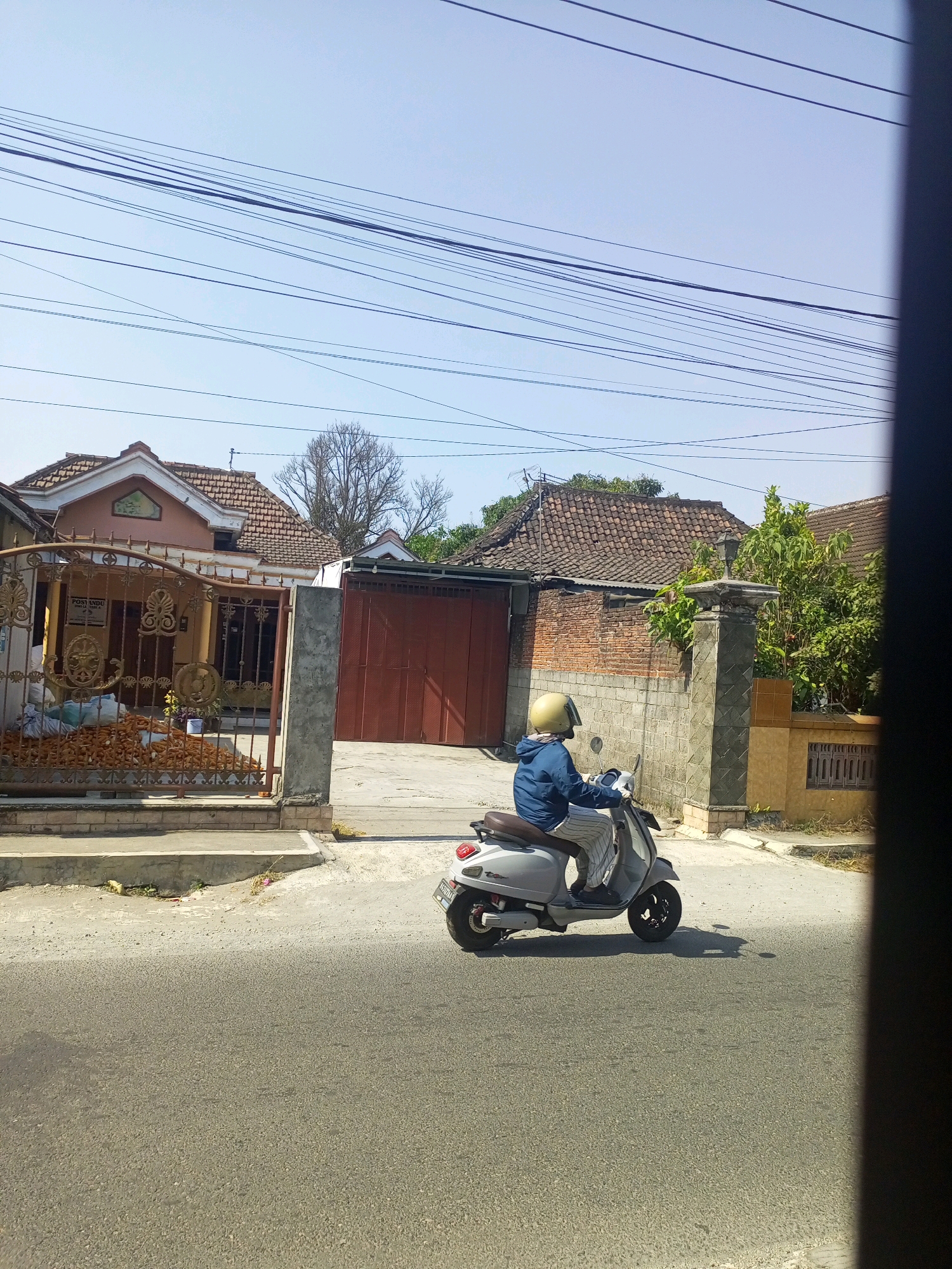 A person wearing a blue jacket and striped pants is riding a light gray scooter with a yellow helmet on a paved road in front of a residential area. The scooter is in motion, casting a shadow. In the background, there are houses with tiled roofs, a brick wall, and a metal gate. Several power lines are visible across the sky. The scene appears to be in a rural or suburban area, likely in Indonesia, given the architectural style and scooter model.