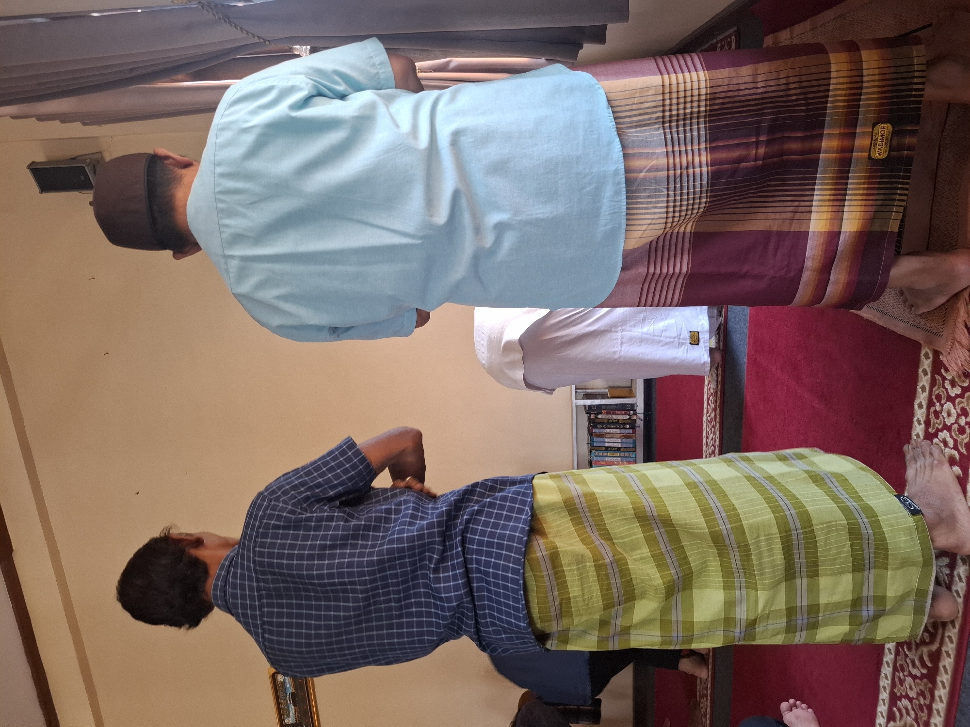 Two men, viewed from behind, stand on a red carpet in what appears to be a prayer room, possibly located in Ciheulang Tonggoh, Indonesia. The man on the left wears a blue and white checkered short-sleeved shirt and a lime green sarong with horizontal and vertical plaid patterns. His arms are crossed behind his back. The man on the right wears a light blue short-sleeved shirt and a dark red and tan sarong with vertical stripes and plaid accents, bearing a "WADIMOR" label. He wears a dark brown cap. To the left of the men, a portion of another person in dark clothing is visible. In the background, a white cloth covers a surface, and a shelf with books is visible. A window with gray curtains is on the right side of the image, and a speaker is mounted on the wall above it. The wall behind the men is a light beige color.
