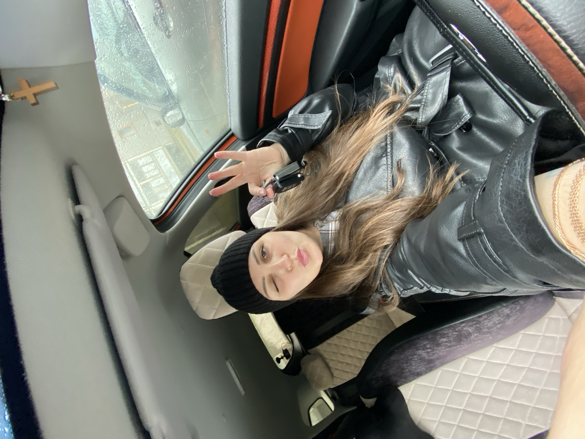 A woman in a car winks at the camera while holding keys and making a peace sign. She wears a black beanie and black leather jacket. It is raining outside. A Christian cross hangs from the car's roof. The car has tan quilted upholstery and orange and black trim. The location is Марковский, Russia.