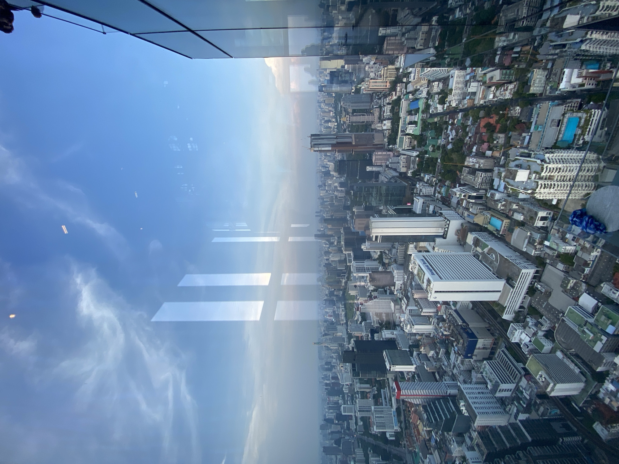 The image depicts a high-angle view of Bangkok, Thailand, as seen through a glass window. The city's skyline stretches into the distance, consisting of numerous buildings, both high-rise and low-rise, interspersed with green spaces indicating trees and vegetation. Some structures include visible swimming pools. The sky is visible both directly and as a reflection in the glass window, showing a mix of blue and light clouds. The bottom section of the window frame is also visible. The scene presents an urban environment with a mix of architectural styles and a hazy atmosphere.