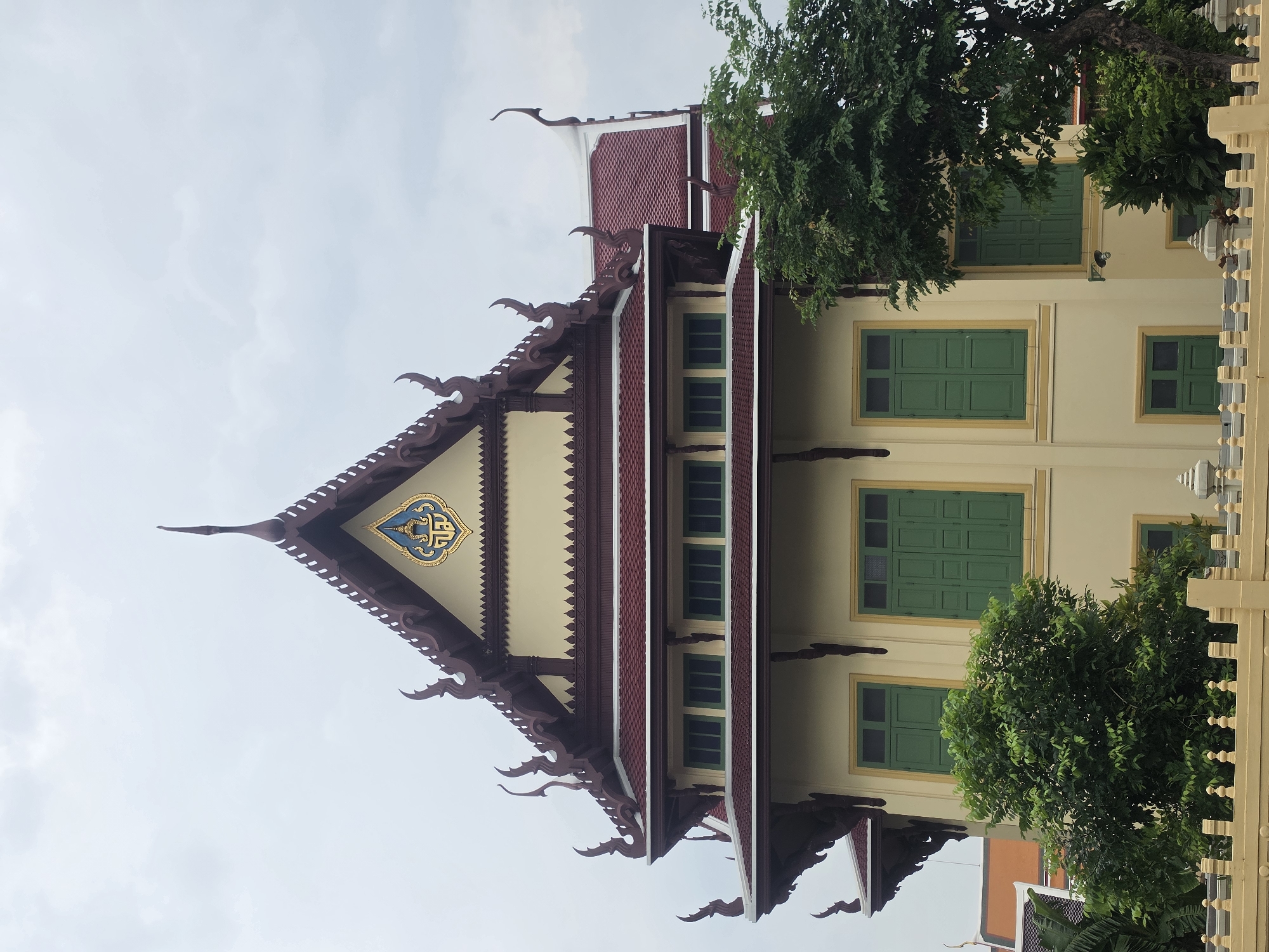 A low-angle, medium shot depicts a building in Bangkok, Thailand. The building's architecture features traditional Thai elements, including a sharply angled, ornamented roof with dark red trim and gold accents. The facade is light yellow with green-framed windows. Foliage partially obscures the building. The sky is overcast.