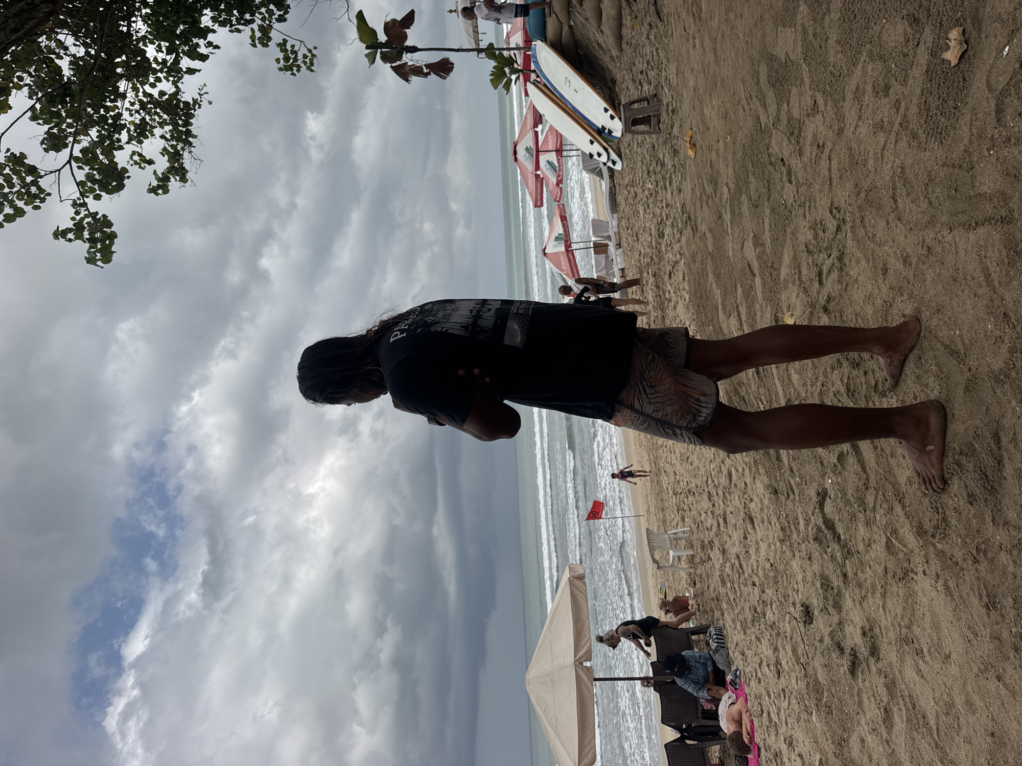This is a medium shot of a beach in Kuta, Indonesia on a cloudy day. A man with long dark hair stands facing away from the camera, with his arms crossed. He is wearing a black t-shirt and patterned shorts. In the background, there are several beach umbrellas, lounge chairs, and people relaxing on the sand. The ocean is visible with waves rolling onto the shore. Two surfboards stand upright on the beach near some red umbrellas. A small red flag is planted in the sand. The overall atmosphere is relaxed and casual.