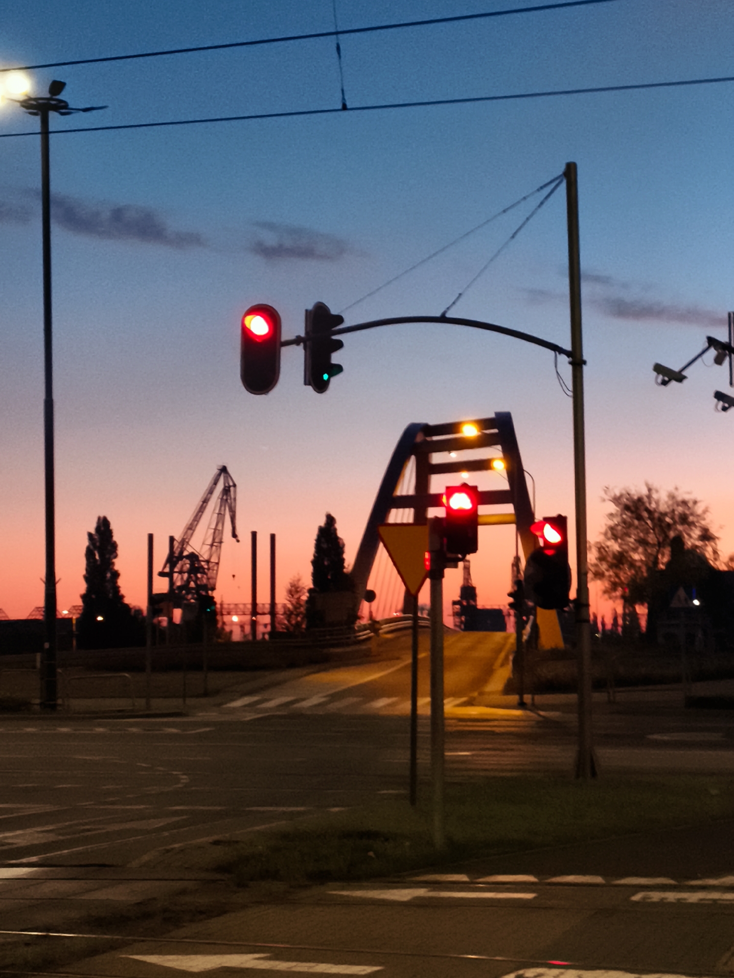 The image captures a scene at dusk in Gdańsk, Poland, likely near the Motława River, given the presence of cranes and industrial elements. The sky displays a gradient of twilight colors, from deep blue at the top to vibrant pink and orange near the horizon. A traffic light with a red signal illuminates the foreground, indicating a stop. In the mid-ground, a bridge structure is visible, with other traffic lights and some yellow warning signs. Further in the background, large industrial cranes are silhouetted against the colorful sky, suggesting a port or shipyard area. Trees and other urban structures are also discernible. The street in the foreground shows road markings, including pedestrian crossings. There are no people clearly visible in the image. The overall impression is of an urban industrial landscape at the end of the day.
