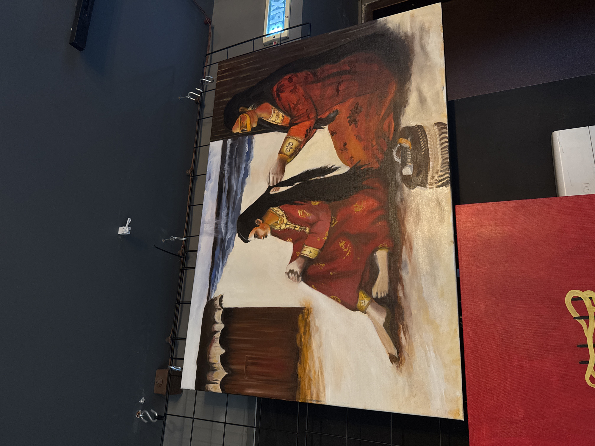 The image displays an oil painting on canvas, captured from a sideways perspective, exhibited in what appears to be an indoor art gallery or exhibition space. The painting depicts two figures, likely women, dressed in elaborate red garments adorned with golden patterns and embroidery. Both figures have long, dark hair. The upper figure in the painting is seen reaching downwards towards the lower figure, suggesting an intimate interaction, possibly grooming or comforting. The lower figure is seated or kneeling, with bare feet visible. The background of the painting features a contrasting scene with broad strokes of white and blue on the left, possibly representing water or sky, and a light, earthy tone for the ground. A dark, wooden-like structure is visible in the foreground of the lower figure.
The painting itself is secured to a black grid display stand, set against a dark grey wall. Above the painting, a light fixture is visible with the Cyrillic word "ВЫХОД" (VYKHOD) prominently displayed, which translates to "EXIT" in Russian. To the immediate right of the main painting, a portion of another red canvas with a swirling golden design is visible. The overall setting indicates an artificial, controlled environment typical of an art show, with various hooks and fixtures on the wall, and what looks like a speaker or a similar device mounted high up. The visible Russian text confirms the given location context of Izhevsk, Russia.