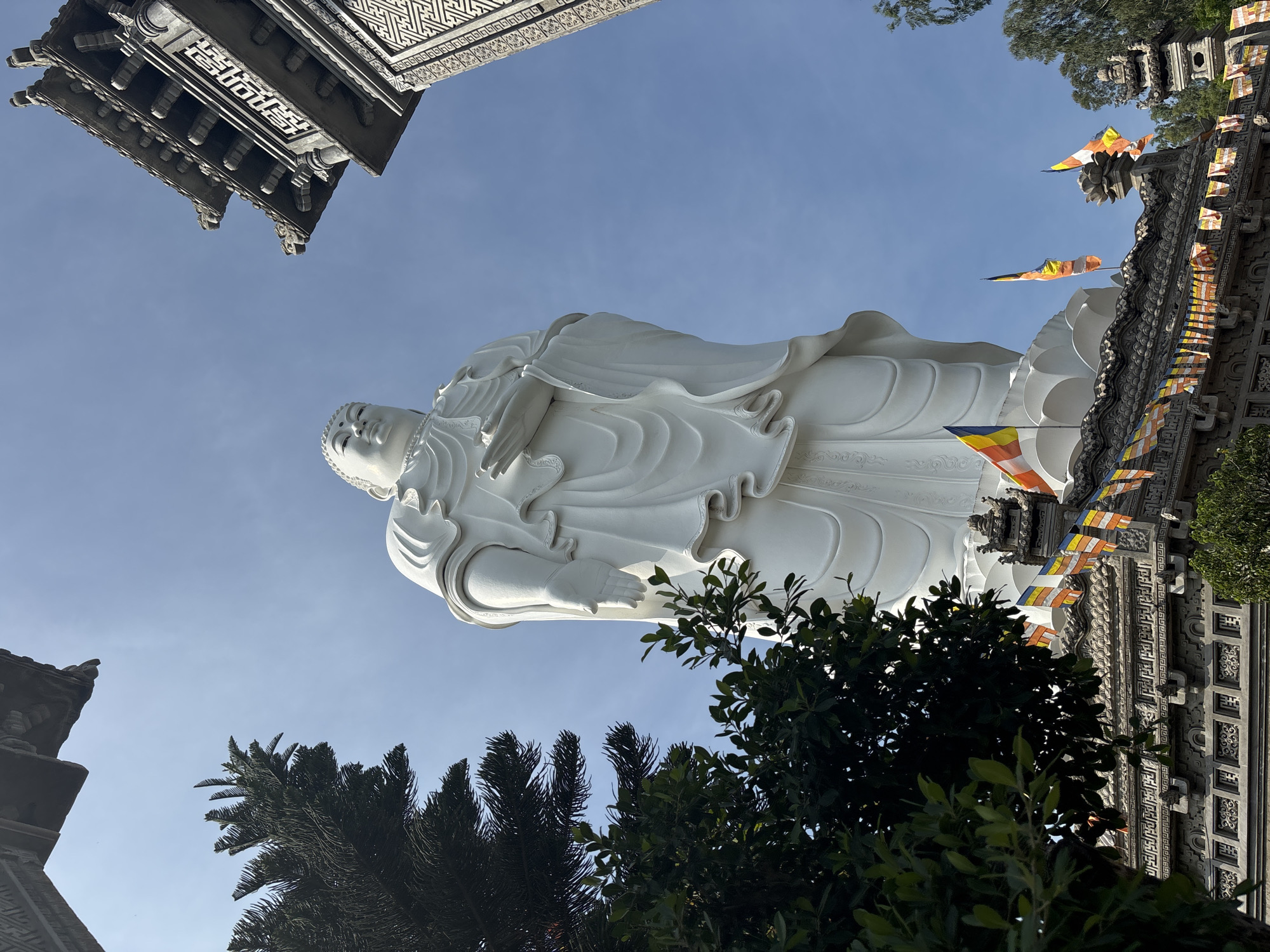 A large, white, serene Buddha statue stands tall against a clear blue sky. The statue is in a standing pose with its hands held together. The base of the statue is adorned with intricate carvings and prayer flags are strung across the platforms below. To the left of the statue, a lush green tree frames the view, and to the right, a portion of a traditional pagoda-style building is visible, also featuring ornate architectural details. The scene appears to be a place of worship or a significant cultural landmark. The location is identified as Tay Nha Trang Ward, Vietnam.