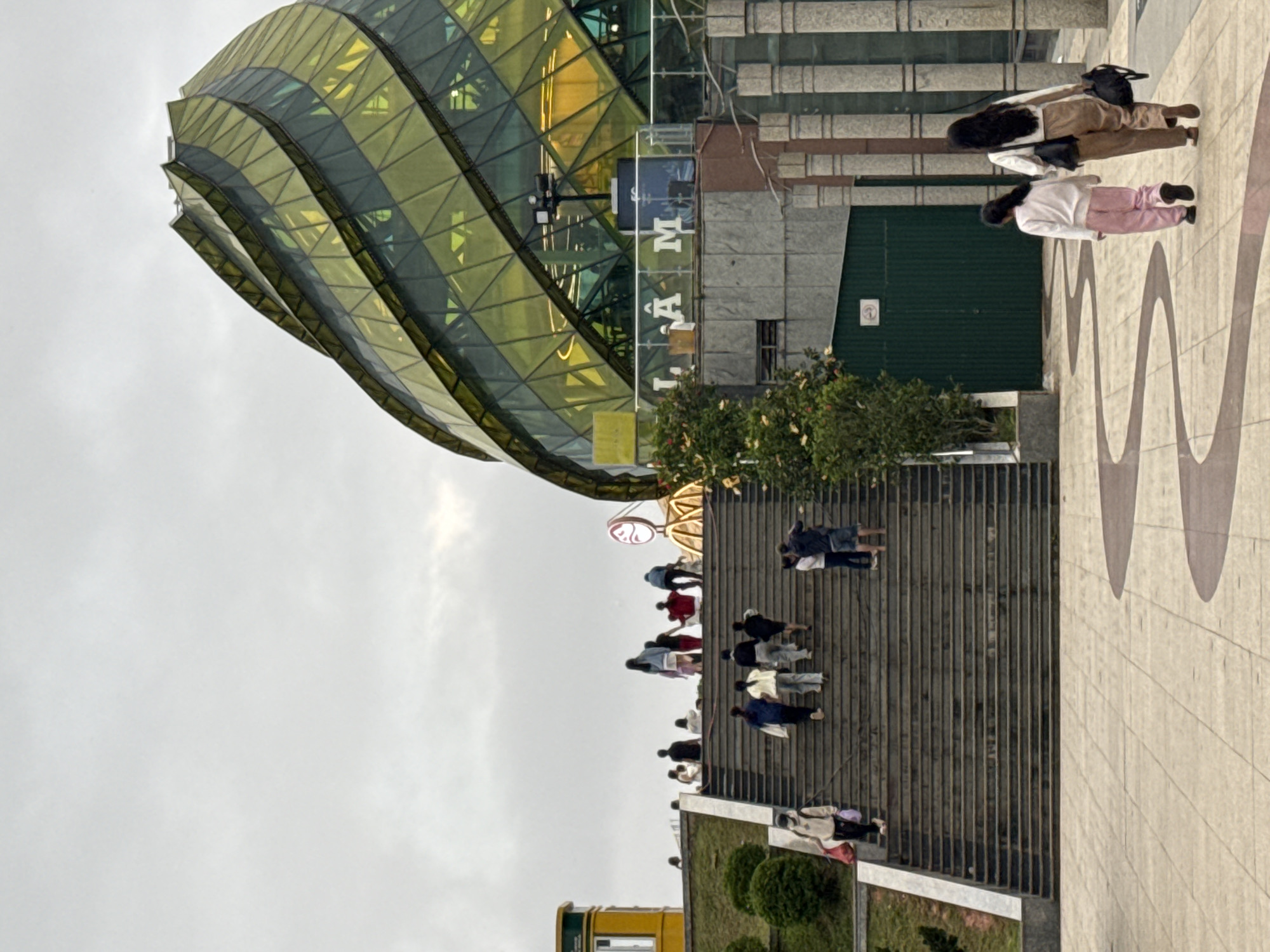The image depicts an outdoor scene in Da Lat, Vietnam, under an overcast sky. The primary subject is a modern building with a distinctive, undulating green and yellow glass facade. In the foreground, a wide, tiered set of concrete steps leads up towards the building. Several people are visible, either walking up or down the stairs, or gathered at the top. Their attire suggests a cool or mild day.
On the ground level near the top of the stairs, two individuals are standing, one wearing a white top and pink pants, and the other a brown jacket and pants, both with dark hair. Further up the stairs, a group of people are ascending. The building itself has a futuristic architectural design, with geometric panels forming its curved structure. Some signage is visible on the building, including letters that appear to spell "LAM."
The scene suggests a public space, possibly a tourist attraction or a shopping center, given the presence of many people and the striking architecture. The lighting is diffused due to the cloudy weather, and the overall mood appears casual and everyday. The presence of trees and greenery around the base of the building adds to the natural setting of Da Lat.