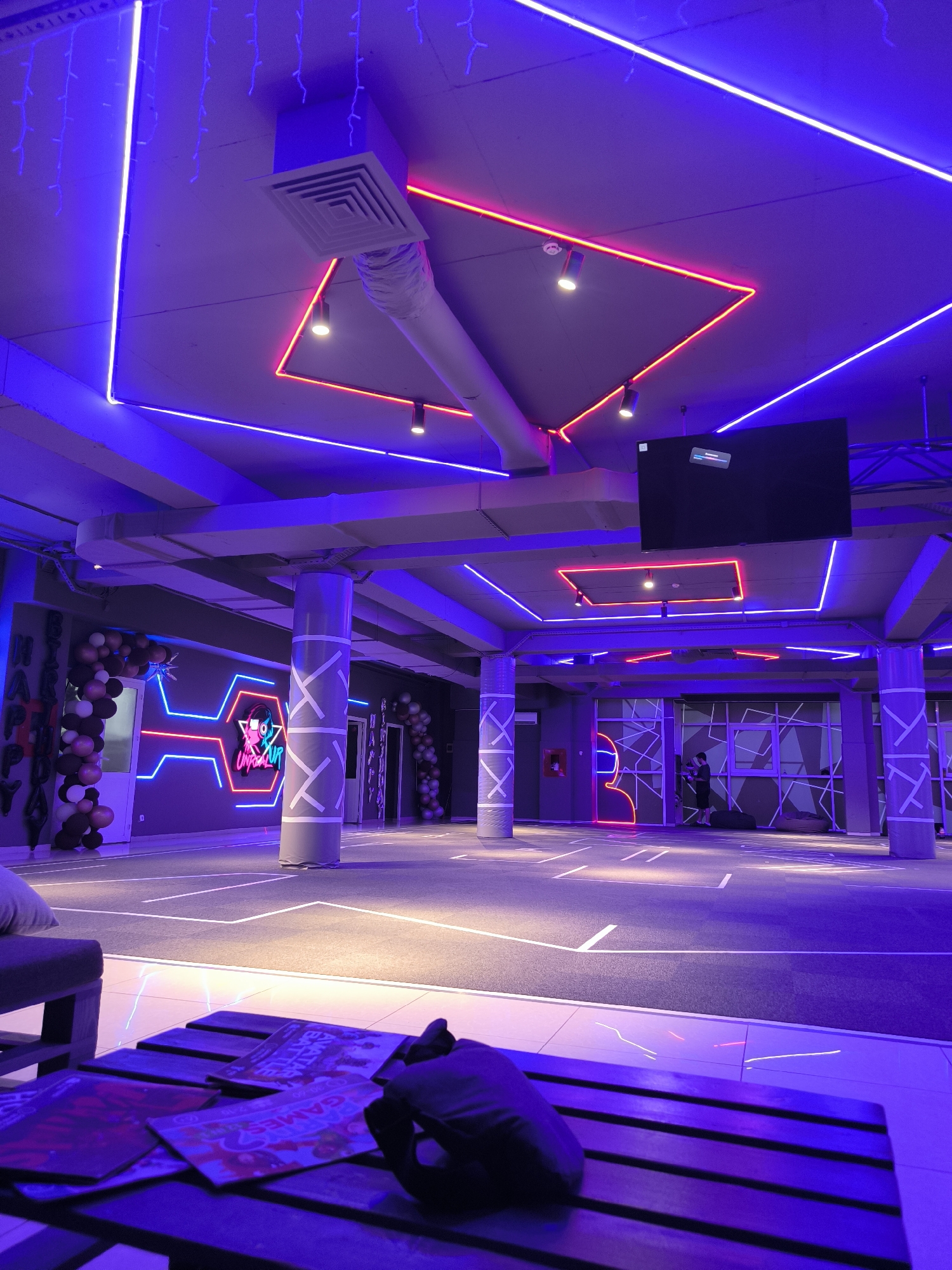 The image shows the interior of a modern entertainment venue, likely a gaming or esports arena, illuminated by vibrant neon lights. The dominant colors are deep blues and purples, with accents of bright pink. The ceiling is adorned with a complex network of blue and pink neon strips, creating geometric patterns. A large black television screen is mounted on the ceiling. The walls are dark gray and feature illuminated neon designs, including a stylized logo with headphones and a humanoid figure. Large cylindrical columns with white criss-cross patterns are spaced throughout the room. The floor is a dark carpet with white lines marking out a playing area. In the foreground, a low wooden table is visible, with several magazines or comic books scattered on its surface, under a purple light. A dark fabric object, possibly a bag or hood, is placed on the table. In the background, a person is visible standing near a wall with geometric window-like designs, and other people are sparsely distributed in the venue. Balloons are arranged in arches on either side of doorways, suggesting a celebration or event. The overall ambiance is futuristic and energetic. This venue is identified as "Battle Arena" in Chișinău, Moldova.