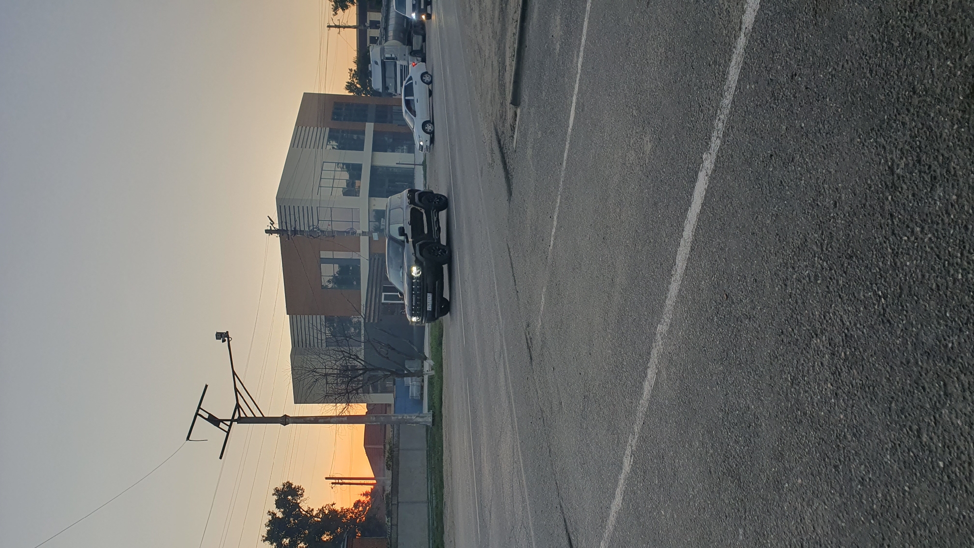 A black SUV is driving on a paved road in Ust-Labinsk, Russia. The sky is clear and orange, indicating it is either sunrise or sunset. In the background, there is a modern building with glass windows and a grey and brown facade. To the left of the building, there are trees and houses. A traffic light pole with a camera is visible on the left side of the image. Other vehicles, including a white car and a truck, are also on the road. The road has white lane markings.