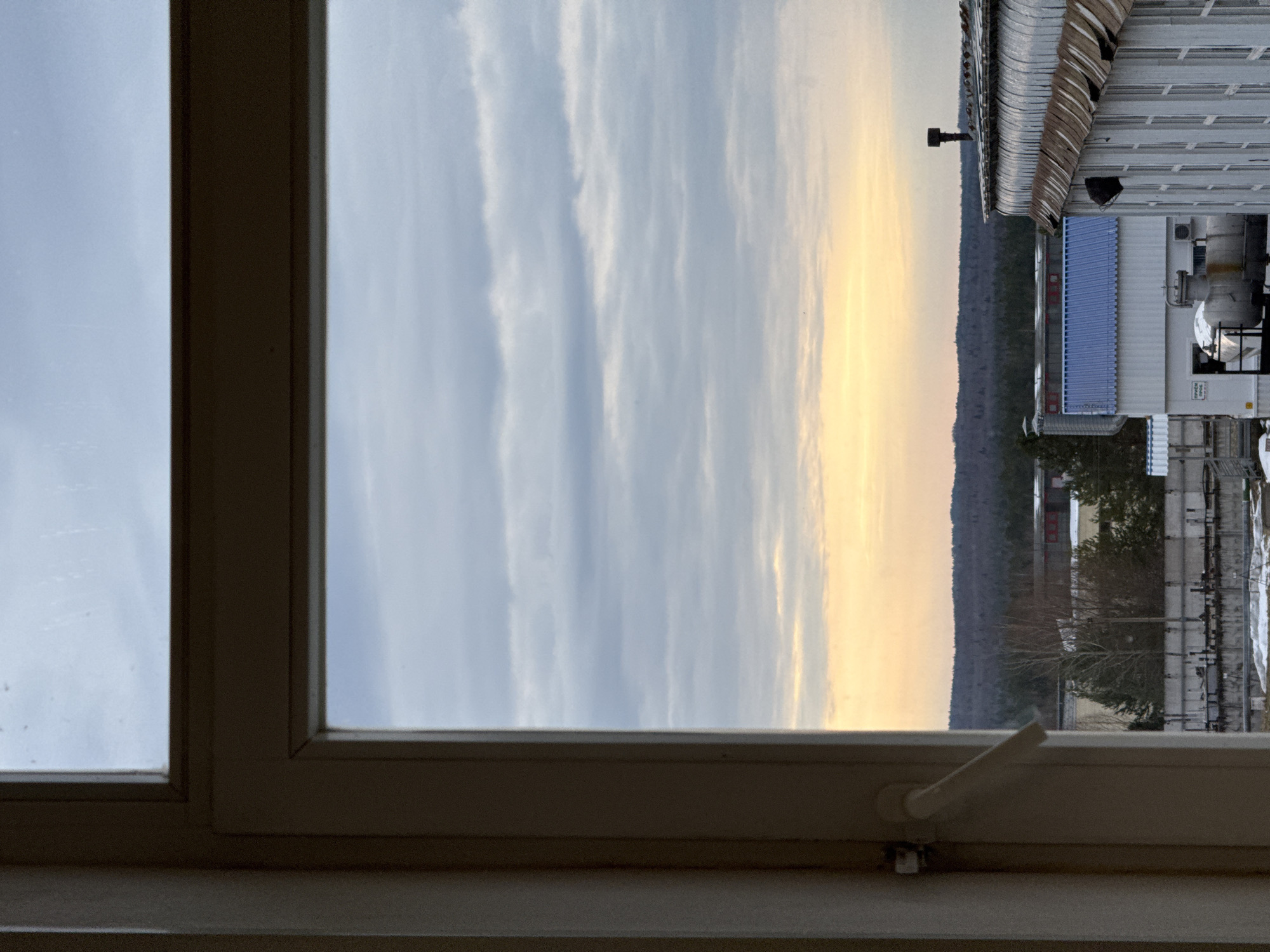 A view from an interior window showing a cityscape during a dawn or dusk period. The sky is visible through a center pane, displaying alternating light blue and pale yellow sections, likely indicative of a sunrise or sunset. Adjacent to the sky are visible elements of an urban industrial area. The setting is possibly Chaikovsky, Russia, based on the industrial elements in the cityscape.