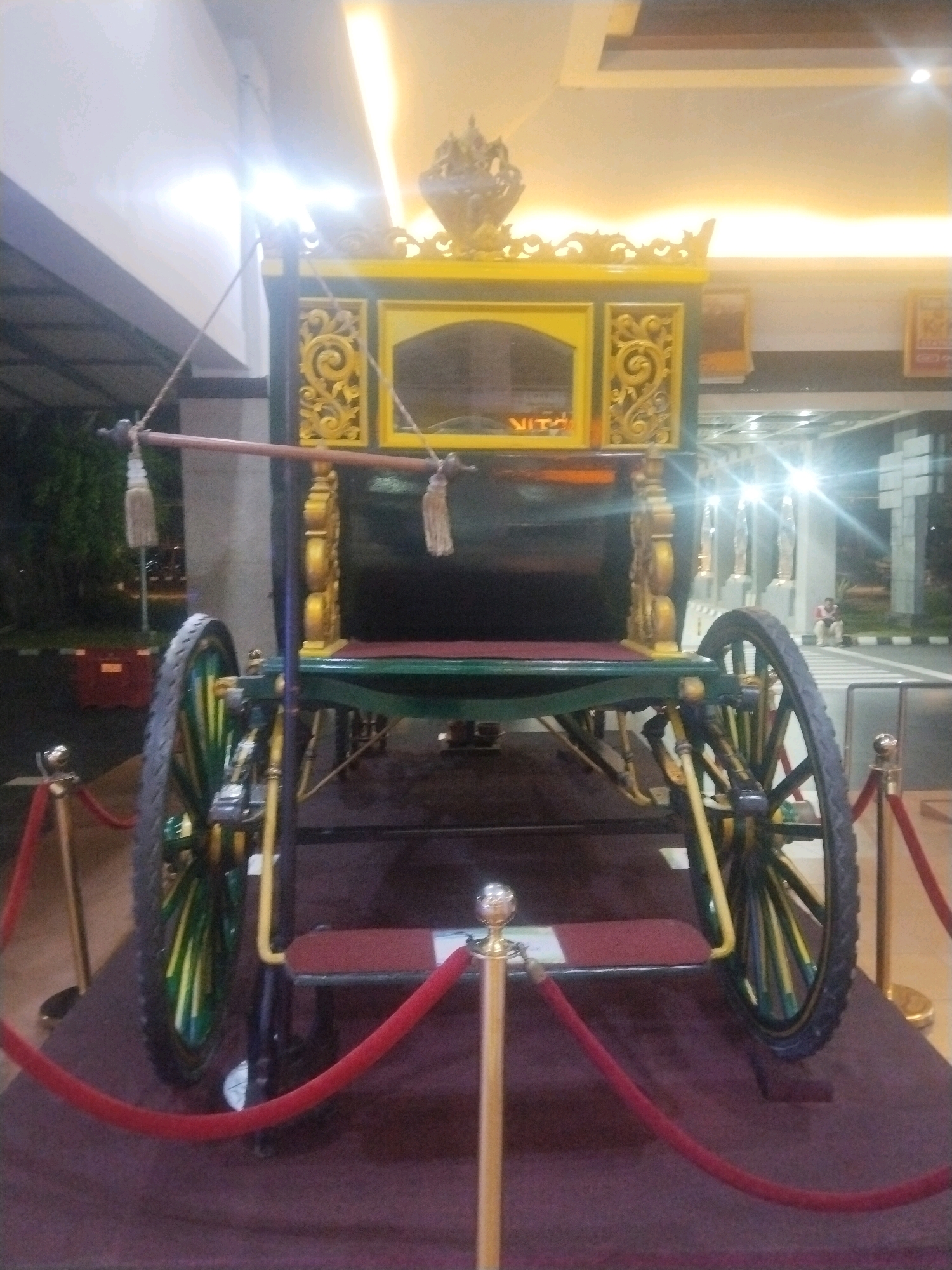 A front-facing, medium-shot photograph captures a decorative, ornate horse-drawn carriage displayed indoors, likely in a public space like a museum or hotel lobby in Desa Ngesrep, Indonesia. The carriage is predominantly green and gold, with intricate carved details on its upper section. A yellow frame surrounds a dark panel on the front of the carriage. The carriage has two large spoked wheels, one on each side, with yellow and green spokes. Red velvet ropes on gold stanchions create a barrier around the carriage, with a deep red carpet extending across the foreground. The background shows a brightly lit interior with what appears to be a building entrance and some greenery outside. A single person is visible in the far background, seated on a bench. The overall lighting is bright, with some glare from overhead lights.