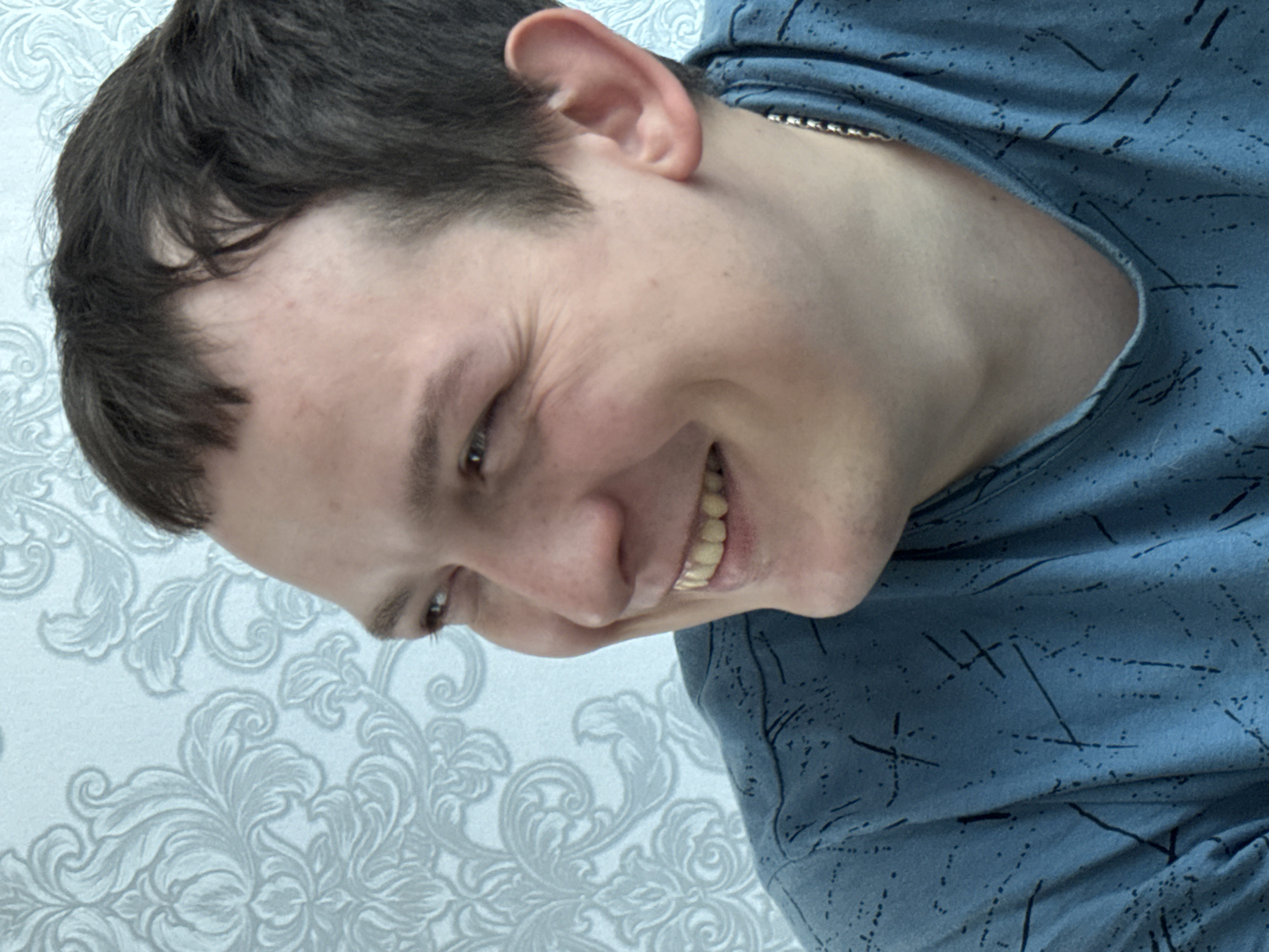 A close-up, tilted shot features a smiling man with dark hair and a light blue t-shirt. The background is a light blue wall with a floral pattern. A glimpse of a chain necklace is visible at the neck of the t-shirt. There is no verifiable evidence to confirm the location is in Кемуль, Russia.