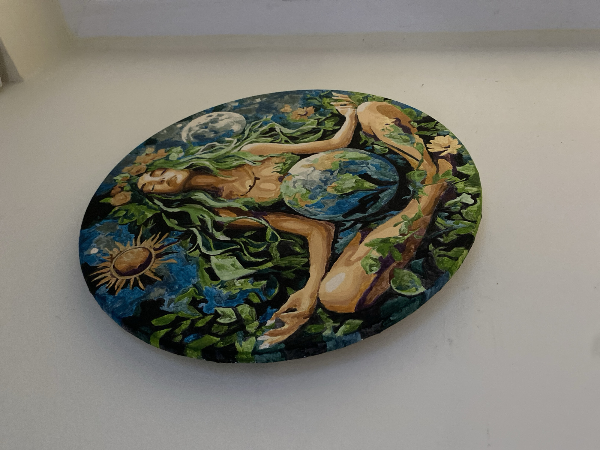 A circular painting depicts Gaia, the Earth goddess, in a meditative pose. She is shown with green, flowing hair, adorned with flowers and leaves. Her eyes are closed, and she cradles a representation of the Earth in her lap. The background features a stylized sun and moon, along with abundant foliage. The painting is mounted on a white wall, angled slightly downwards, suggesting it is hanging in a room. The lighting appears to be from an overhead source. The painting is located in Chornomorsk, Ukraine.