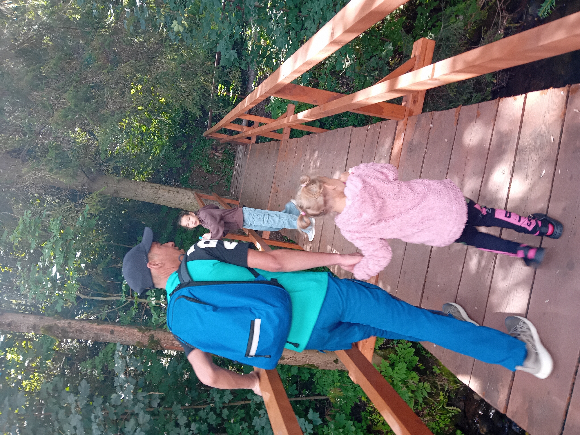 In Zuberec, Slovakia, a man wearing a blue backpack and a turquoise shirt with black accents, along with blue pants and a black cap, is walking across a wooden bridge. He is holding the hand of a young girl in a pink furry jacket and black leggings with pink accents. Behind them, a girl with dark hair pulled back in a ponytail, wearing a brown hoodie and blue jeans, is also on the bridge. The bridge is surrounded by lush green trees and foliage, indicating an outdoor, natural setting. Sunlight filters through the leaves, casting dappled shadows on the wooden planks of the bridge. A small stream is visible below the bridge on the left side of the image.