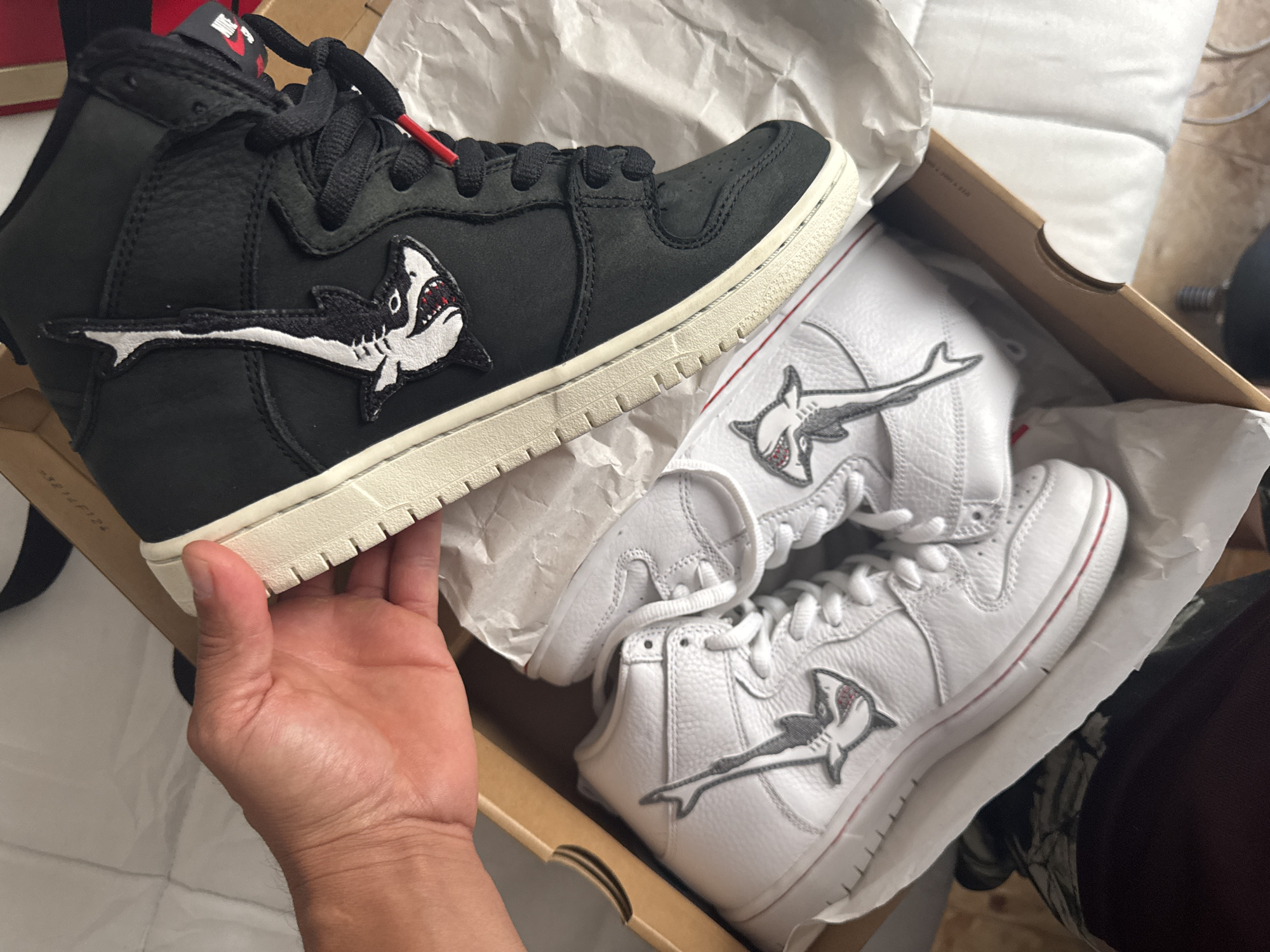 The image shows a pair of sneakers inside a shoebox, held in a hand. The shoebox is tan and lined with white tissue paper. One sneaker is black with a white sole and a shark patch, held in hand. Inside the box are two white sneakers with a shark patch. The location of the image is not discernable from the given image.