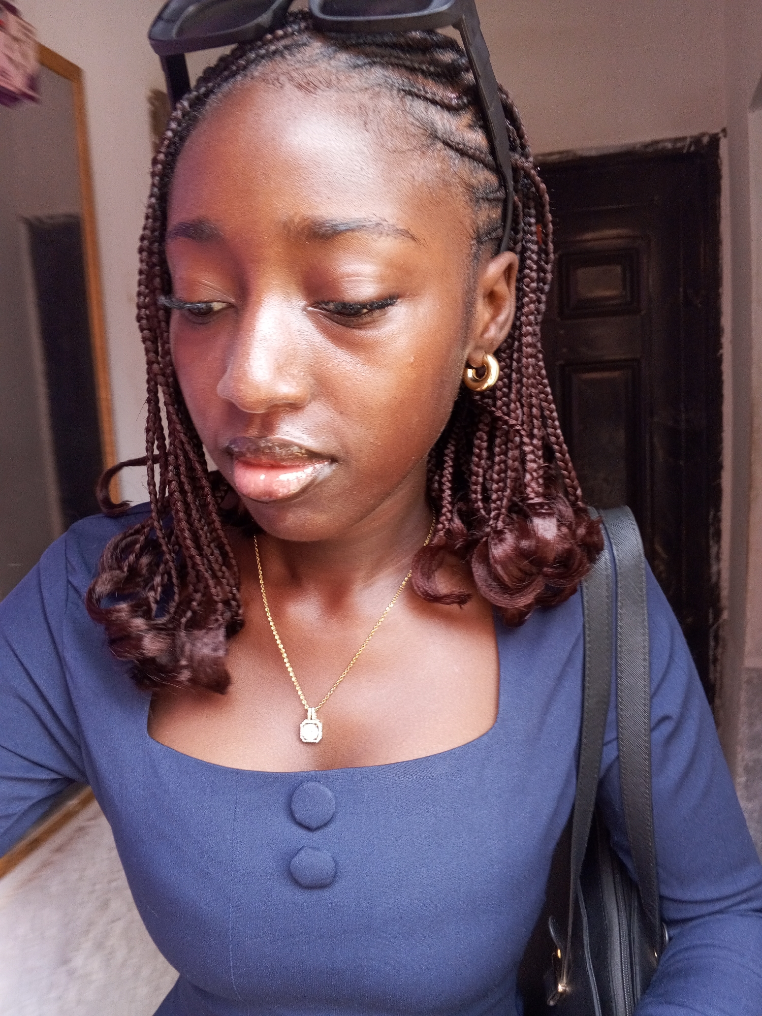 A medium shot captures a young, dark-skinned woman with braided hair, accessorized with sunglasses on her head and gold hoop earrings. She's wearing a navy blue dress with two buttons and a delicate gold necklace. A black shoulder bag hangs from her shoulder. Her gaze is directed downward. The setting includes a partly visible mirror and a dark brown door, suggesting an indoor environment. The location is likely in a residential or commercial area in Abuja, Nigeria, based on the architectural style and typical indoor aesthetics.