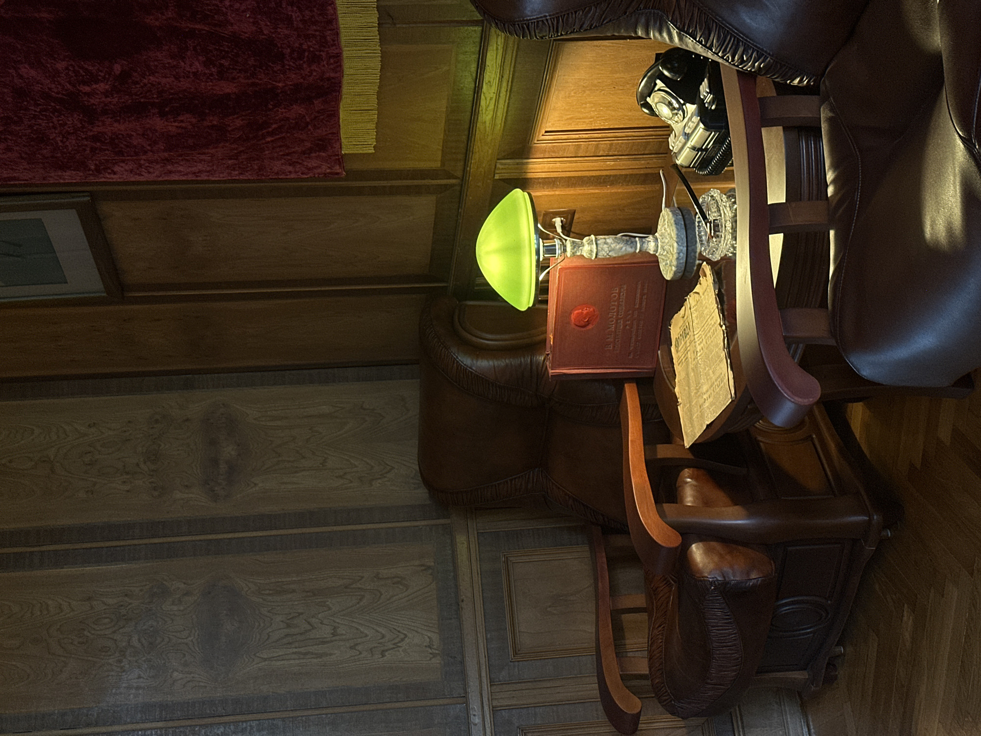 The image presents a scene indoors, likely a study or office space. The setting is characterized by rich, dark wood paneling on the walls and a herringbone-patterned wood floor.
Prominent features include a large, plush leather armchair, positioned near a small wooden table. A rotary telephone is placed on the back of the chair. On the table, there is a green-shaded lamp, a book, and some papers or documents. The book appears to have Russian text on its cover. A small carpet with a maroon and yellow fringe is visible on the wall.
Given the presence of the book with Russian text and the style of the room, it is plausible this location is in Sochi, Russia, or another area where Russian cultural elements are prevalent.