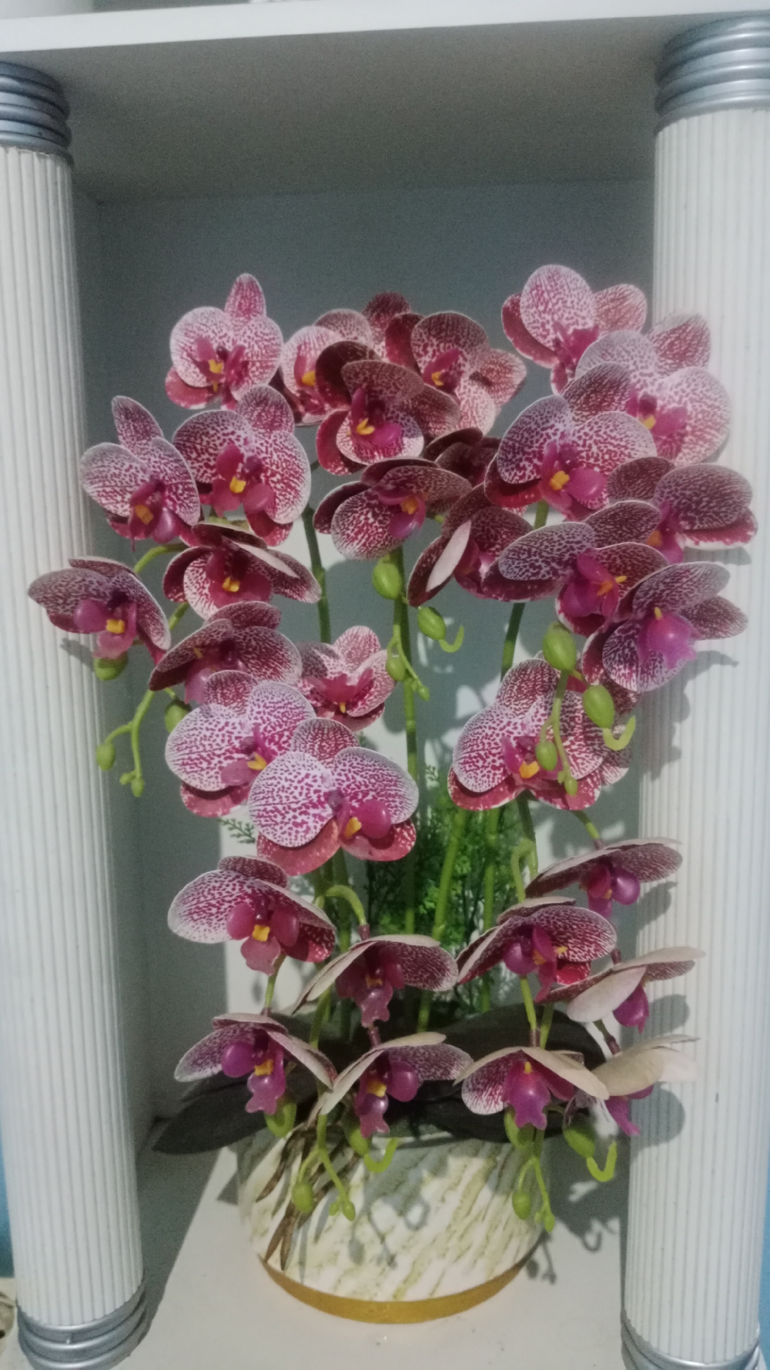 A vertical, medium shot captures an arrangement of artificial purple and white spotted orchids in a decorative pot, flanked by two fluted white columns. The orchids are densely packed on multiple stems, with some flowers open and others in bud. The pot has a light-colored, textured pattern with a golden rim and sits on a white surface. The background is a plain light gray. The scene appears to be indoors, possibly in a display or home setting. There are no people visible.