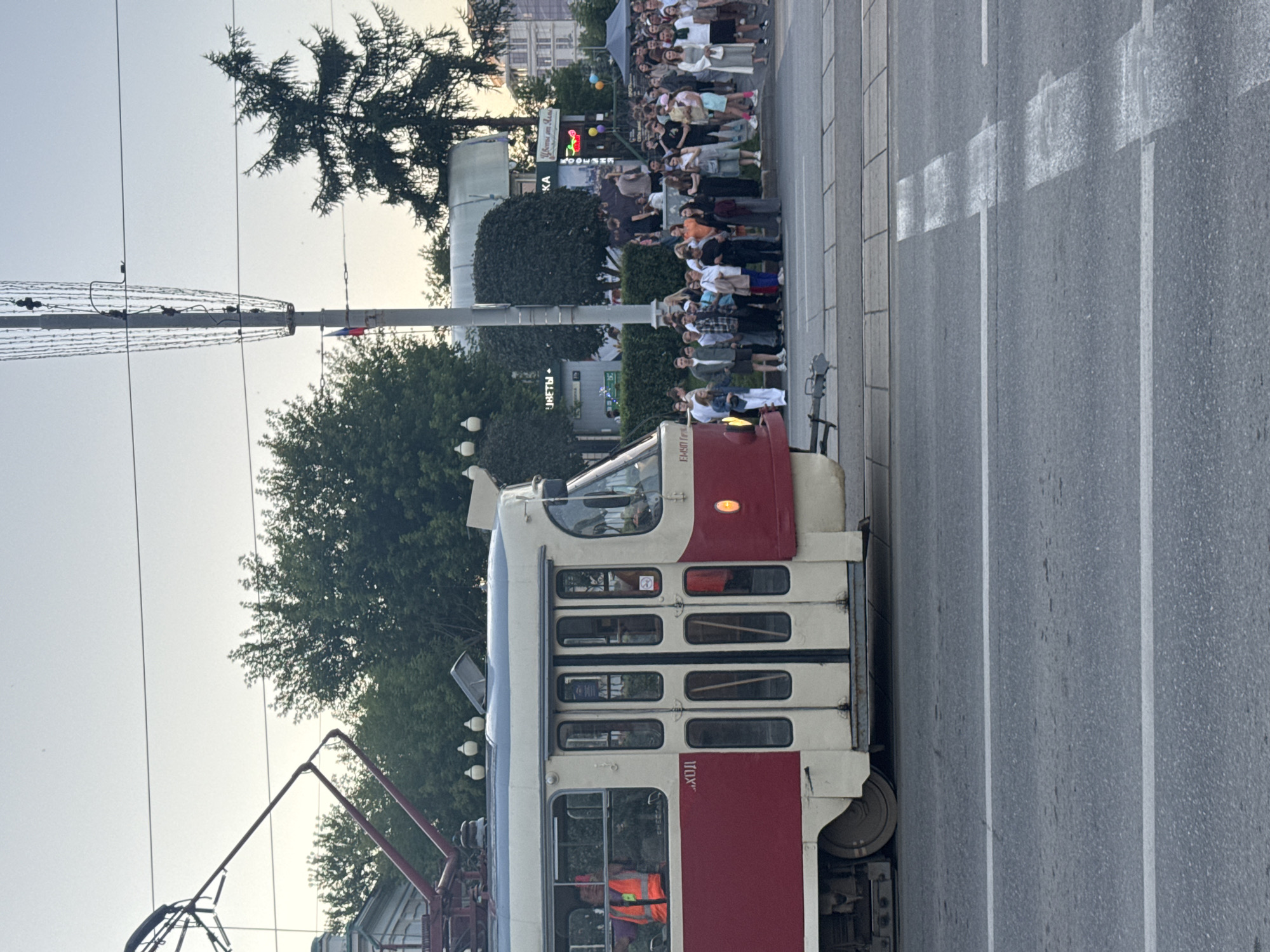 This image captures a scene on a city street in Yekaterinburg, Russia, likely near the Yekaterinburg City Administration building, given the architecture and the presence of public transportation. A red and cream-colored tram is stopped at a streetcar stop, with its doors open. A driver wearing a high-visibility orange vest is visible through the front window.
A large group of people, appearing to be diverse in age and gender, are gathered on the sidewalk, waiting. Some are standing in a queue, suggesting they are waiting to board the tram. The background reveals trees, streetlights, and buildings, including a building with a sign that reads "ЦВЕТЫ ОТ МАМА" (Flowers from Mama) and another partially visible sign with Russian text. The sky is overcast with a soft, diffused light, indicating it is likely early morning or late afternoon. The street in the foreground has white lane markings. The overall atmosphere appears to be calm and ordinary, depicting a typical urban street scene.