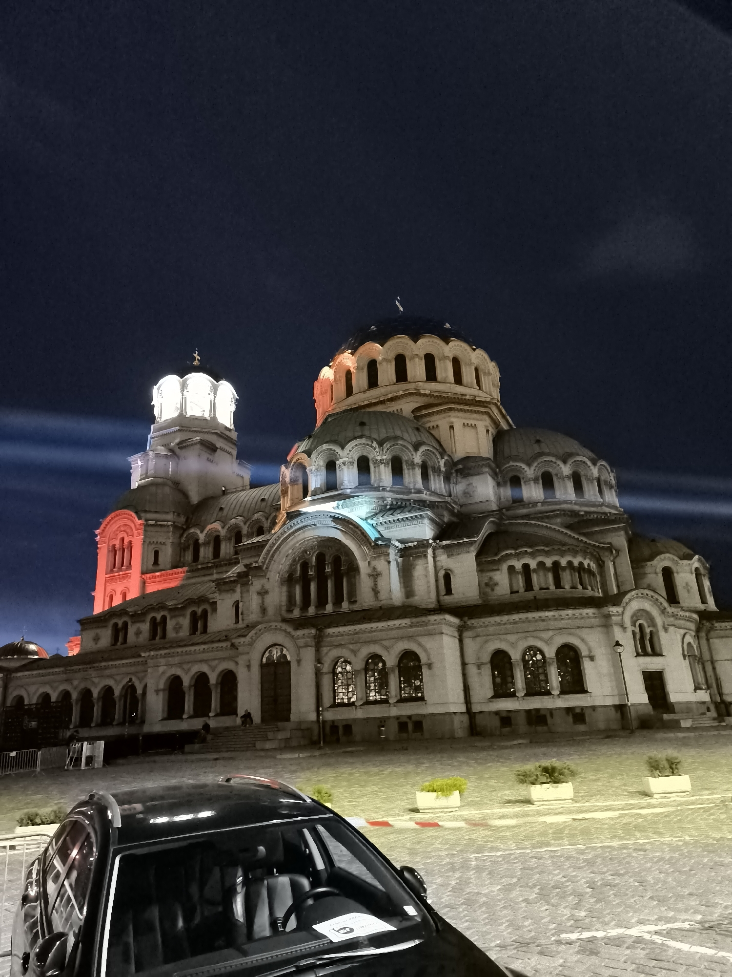 The image captures the Alexander Nevsky Cathedral in Sofia, Bulgaria, at night. The grand cathedral, illuminated by various colored lights, dominates the frame with its numerous domes and intricate architecture. The sky above is dark, with subtle hints of clouds and light beams emanating from the cathedral complex. In the foreground, the front of a black SUV is visible, parked on a cobblestone street. The interior of the car, including the steering wheel and seats, is partially visible through the windshield. The scene is illuminated, suggesting it is either dusk or night, with artificial lighting highlighting the cathedral and surrounding area. There are a few potted plants visible in front of the cathedral, adding a touch of greenery to the scene. A fence is also visible to the left of the cathedral, and some people can be seen in the distance on the steps of the cathedral.