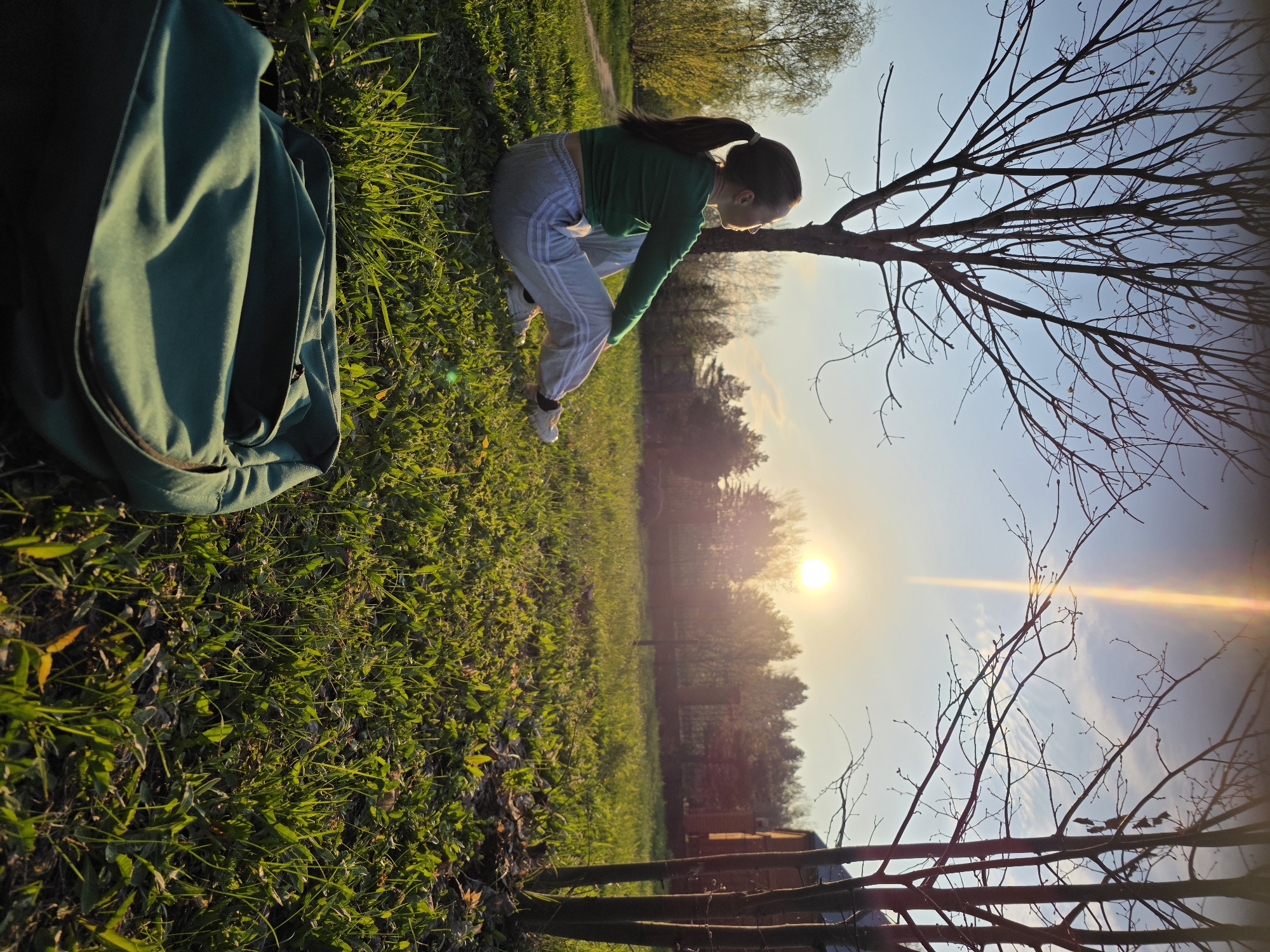 A full shot captures a person seated in a grassy area, likely a park, in Chaikovsky, Russia. The individual, with long dark hair pulled back in a ponytail, is wearing a green long-sleeved shirt, and light grey sweatpants with white stripes. They are in a crouched position, looking towards the ground. To the left of the frame, a large, dark green backpack sits on the grass. The background reveals a sunset over a wooded area, with a distinct sun flare. Bare branches of a tree are visible near the person. Fencing is visible in the lower right corner.