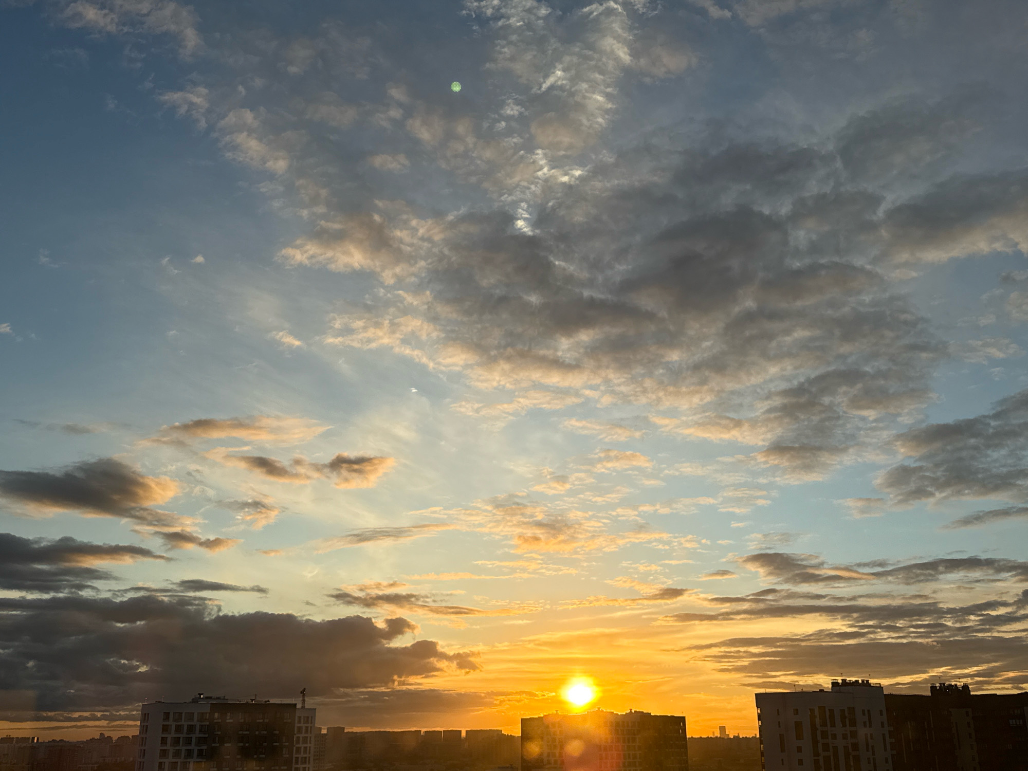 The image depicts a sunset over the city of Lyubertsy, Russia. The sky is filled with a mix of light blue and dramatic orange and yellow clouds, with the sun positioned low on the horizon, casting a bright glow. Silhouetted against the vibrant sky are several multi-story buildings, appearing as dark shapes with visible windows. Some buildings have flat roofs with visible architectural details. The overall scene is one of an urban environment at dusk, characterized by the interplay of natural light and man-made structures.