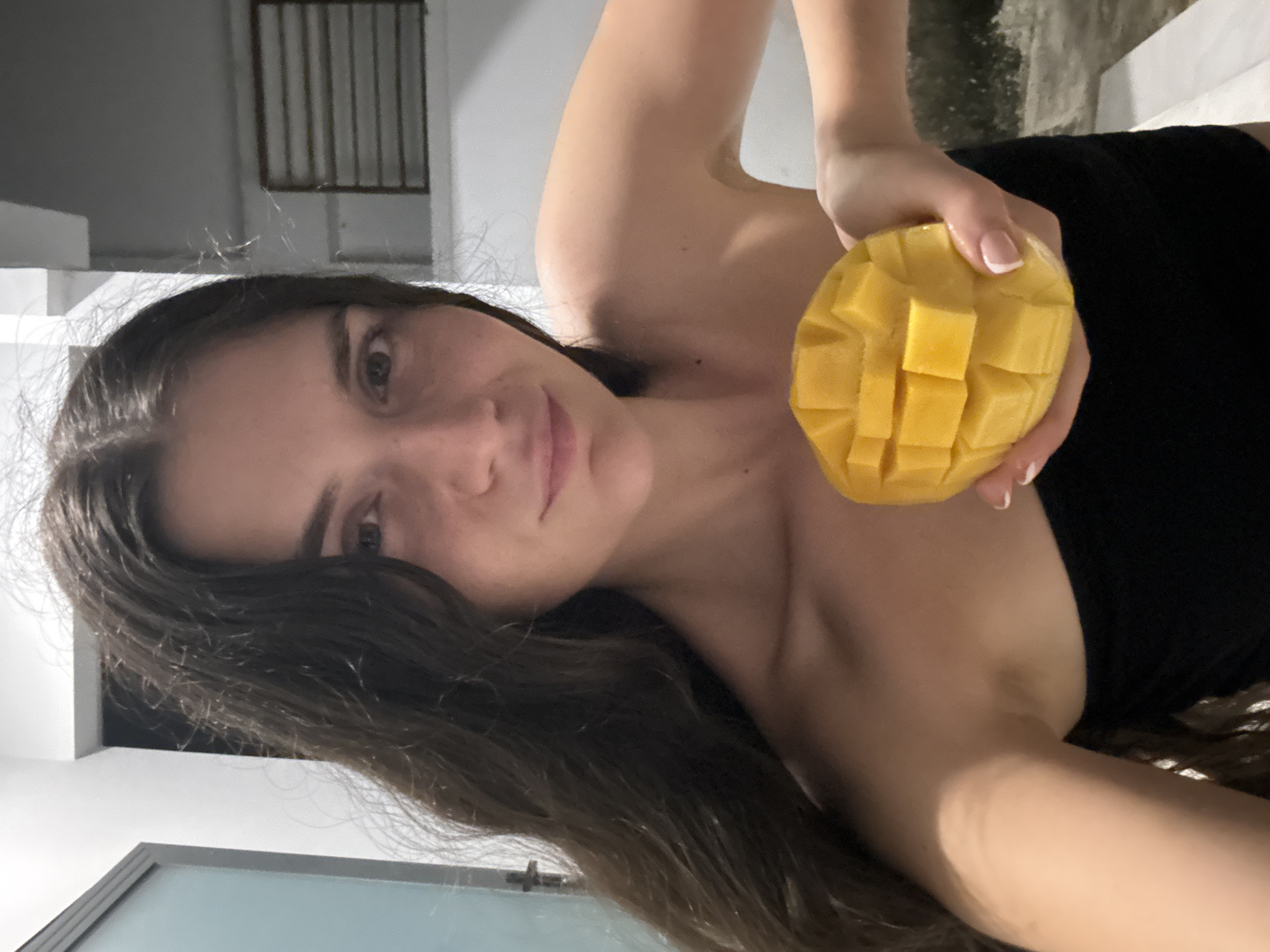 A young woman is holding a peeled mango close to the camera. She has long brown hair, fair skin, and dark eyes. She is wearing a black top. The mango is cut into cubes. The background appears to be a building with a barred window, possibly in Mũi Né, Vietnam. The image is lit with natural light.