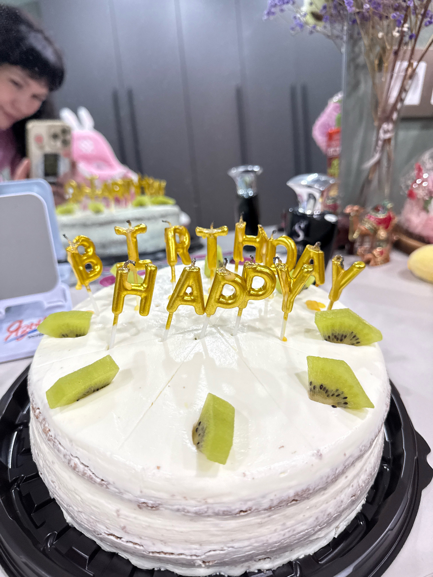 The image shows a birthday cake decorated with gold "HAPPY BIRTHDAY" candles and kiwi slices. The cake is on a table, and in the background, there are other decorative items and what appears to be a blurred reflection of a person. The scene is likely indoors, possibly in a home or a party venue. There is no information in the image that indicates the location is Cheboksary, Russia.