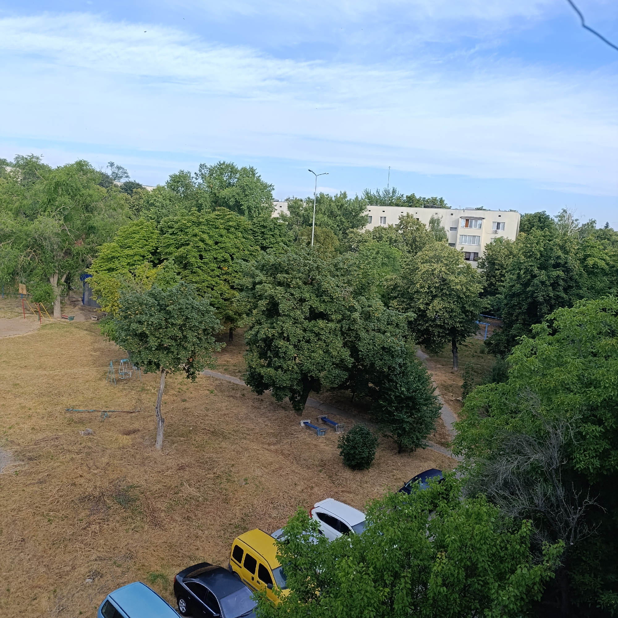 The image depicts an aerial view of a residential area in Ladyzhyn, Ukraine, on a clear day with a partly cloudy sky. In the foreground, dry, yellowed grass covers the ground, with several parked cars visible. These include a light blue sedan, a black sedan, a yellow minivan, and a white hatchback. Some trees with lush green foliage are scattered throughout the scene, and a playground structure with swings and a teeter-totter is also present. In the background, apartment buildings with white facades and visible windows are nestled amongst dense green trees. Streetlights are also present in the background. The overall impression is that of a typical urban or suburban neighborhood.