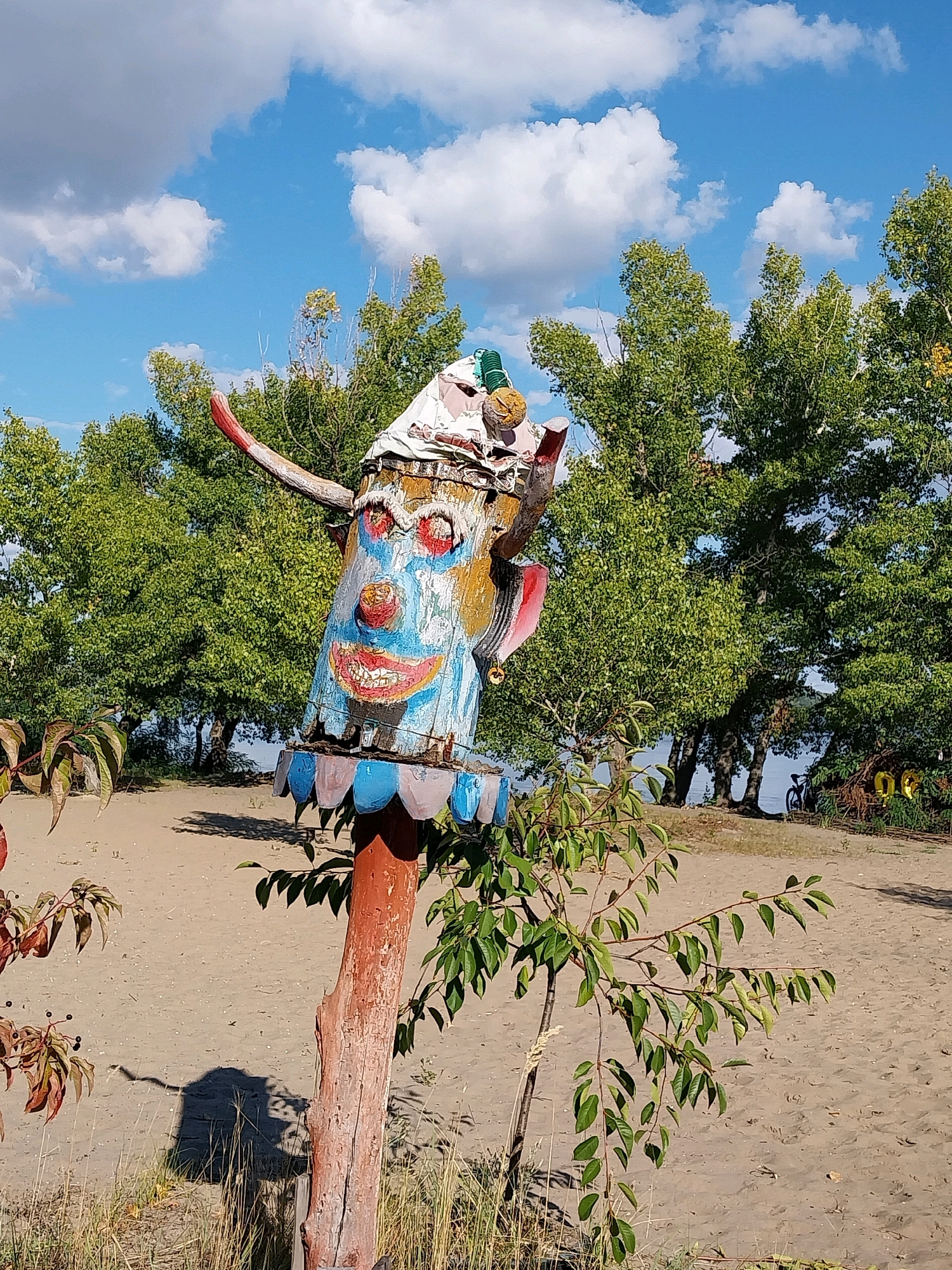 The image depicts a colorful, hand-painted wooden totem pole on a sandy beach. The totem pole is shaped like a clown's face, with exaggerated features such as large red eyes, a blue nose, and a wide, grinning mouth with white teeth. It is adorned with red, blue, and yellow paint. A single red horn protrudes from the left side of its head. The totem pole is mounted on a reddish-brown wooden pole, with a frilly blue and white base around its neck.
In the background, lush green trees line the shore of a body of water, possibly a river or lake. The sky is bright blue with scattered white clouds. A path or road runs behind the totem pole, where a bicycle is parked near some decorations. The ground is covered in sand and sparse vegetation. The overall setting appears to be an outdoor recreational area.
This image was taken in the city of Dnipro, Ukraine, specifically along the Dnipro River embankment.