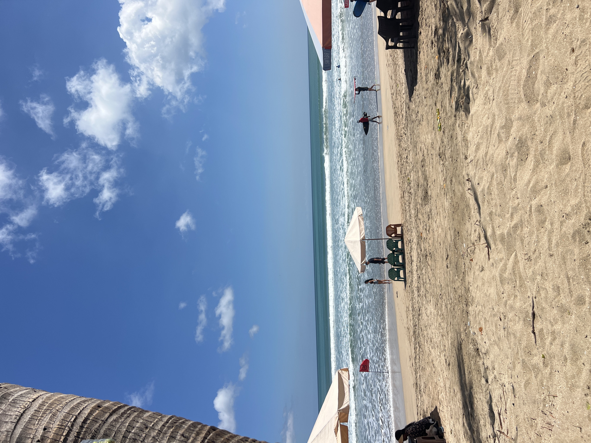 This image depicts a lively beach scene in Kuta, Indonesia, on a bright, sunny day with scattered clouds. The foreground is dominated by textured sand, with sparse debris scattered across its surface. In the midground, the sandy shore meets the gentle waves of the ocean, marked by white foam.
Several people are present:
* On the left, a person wearing a dark hat and dark clothing is seated.
* Near the water's edge, two individuals are standing and looking towards the ocean.
* Further to the right, two people are walking along the shoreline, each carrying a surfboard. The person on the left is wearing a red top and black shorts, while the person on the right is wearing black shorts.
* In the distance, other figures are visible in the water, possibly swimming or surfing.
Various objects enhance the beach atmosphere:
* Two large beach umbrellas are set up, one on the far left and another with an orange and white striped canopy on the right, casting shadows on the sand.
* Beneath the umbrella on the right, four plastic chairs (two green, two brown) are arranged.
* A red flag is planted in the sand near the water, serving as a marker.
* Two surfboards are visible in the sand near the chairs.
* A few small, yellow objects are scattered on the sand between the chairs and the water.
The environment is characterized by:
* A clear blue sky with fluffy white clouds.
* The turquoise ocean with rolling waves.
* The sandy beach typical of Kuta.
* The trunk of a palm tree is visible on the far left, framing the scene.
The overall impression is of a typical, active beach day in Kuta, with people enjoying the sun, sand, and surf.