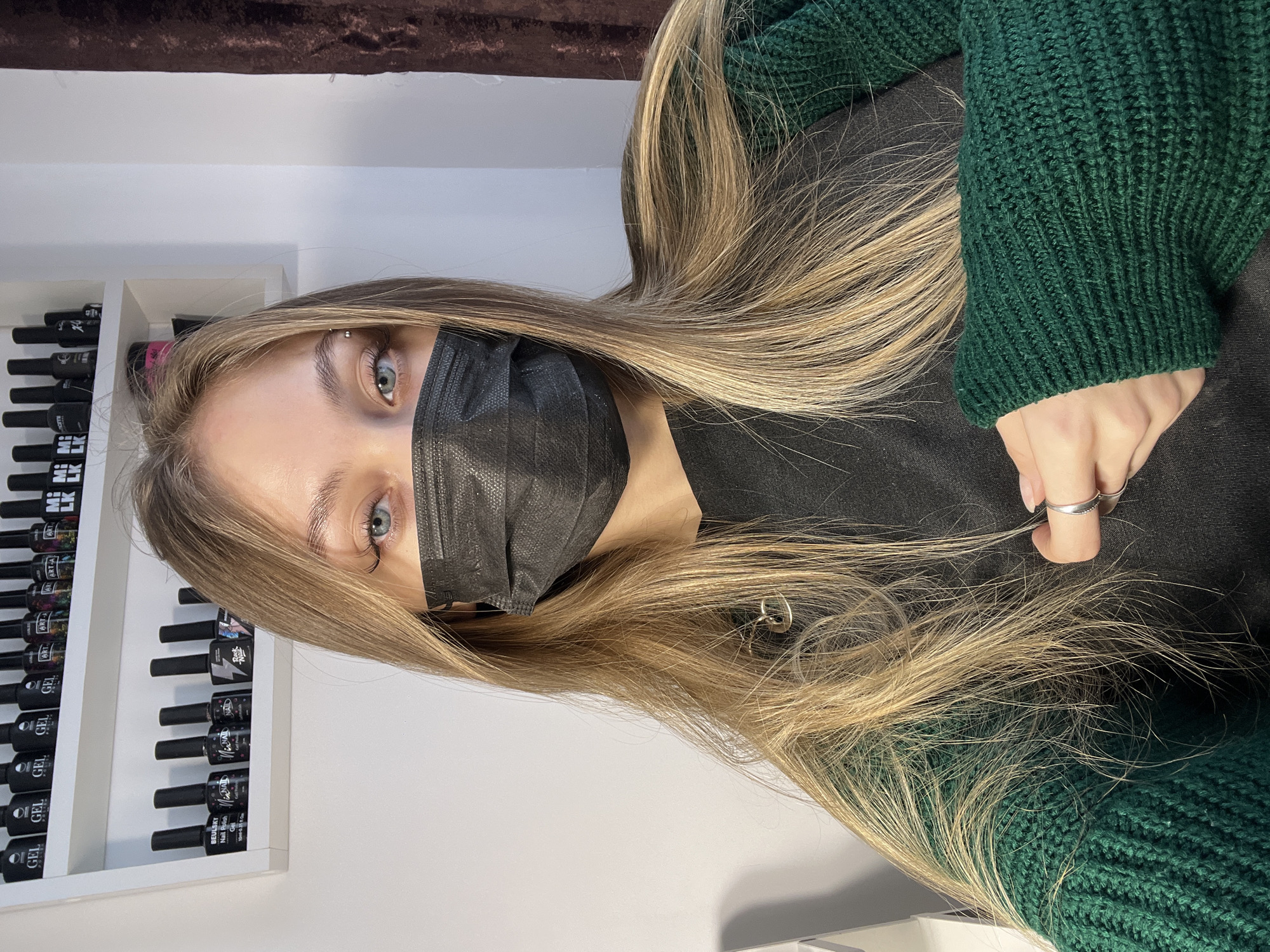 A young woman wearing a black face mask and a dark green knit sweater is the central subject of the image. Her long, blond hair falls over her shoulders and chest. A silver ring is visible on her finger. To her left, a white shelf holds rows of black nail polish bottles. The background includes a white wall and a brown curtain. The location is possibly in the city of Фоки, Russia.