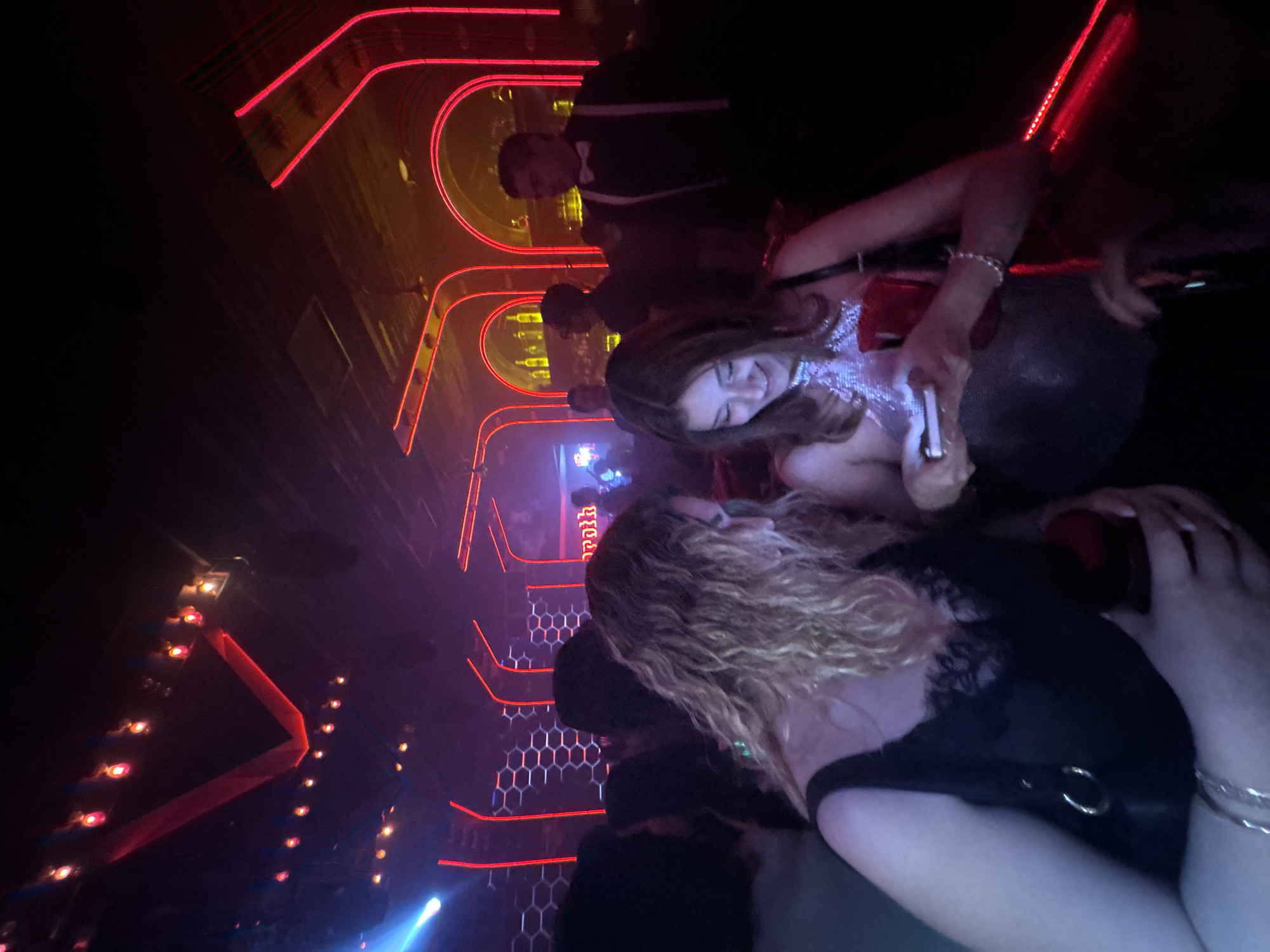 In a nightclub in Puebla, Mexico, two women are in the foreground, one with blonde curly hair and wearing a black top with lace detail, holding a red drink in her right hand. The second woman is wearing a sparkling silver top and looking down at her phone, which she is holding in her right hand. Behind them, a man in a tuxedo stands in a crowded, dimly lit space. The nightclub features red neon lighting in arch and geometric designs. Other figures are visible in the background.