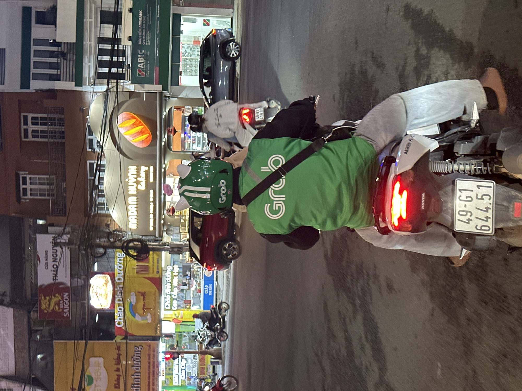 The image depicts a street scene at night in Phường Xuân Hương - Đà Lạt, Vietnam. The primary subjects are two individuals on a motorcycle. One person is wearing a bright green Grab jacket and helmet, suggesting they are a delivery driver. The other person, also on the motorcycle, is wearing light grey pants and a dark jacket.
The motorcycle itself is visible, with a license plate reading "49-G1 644.51". The scene is set along a street lined with shops and buildings. Several signs with Vietnamese text are prominent, including "SAIGON", "MADAM HUỲNH Bakery", and various advertisements for "Cháo Dinh Dưỡng" (Nutritious Porridge). One sign indicates "BẢO HIỂM AGRIBANK" (Agribank Insurance). The presence of streetlights and illuminated shop signs confirms it is nighttime.
The overall atmosphere appears to be a typical urban evening in Vietnam, with the flow of traffic and commercial activity. There are no overt emotional cues, but the subjects are focused on their journey or task. The weather appears to be clear, as there are no visible signs of rain. The image is slightly tilted, adding a dynamic perspective to the street view.