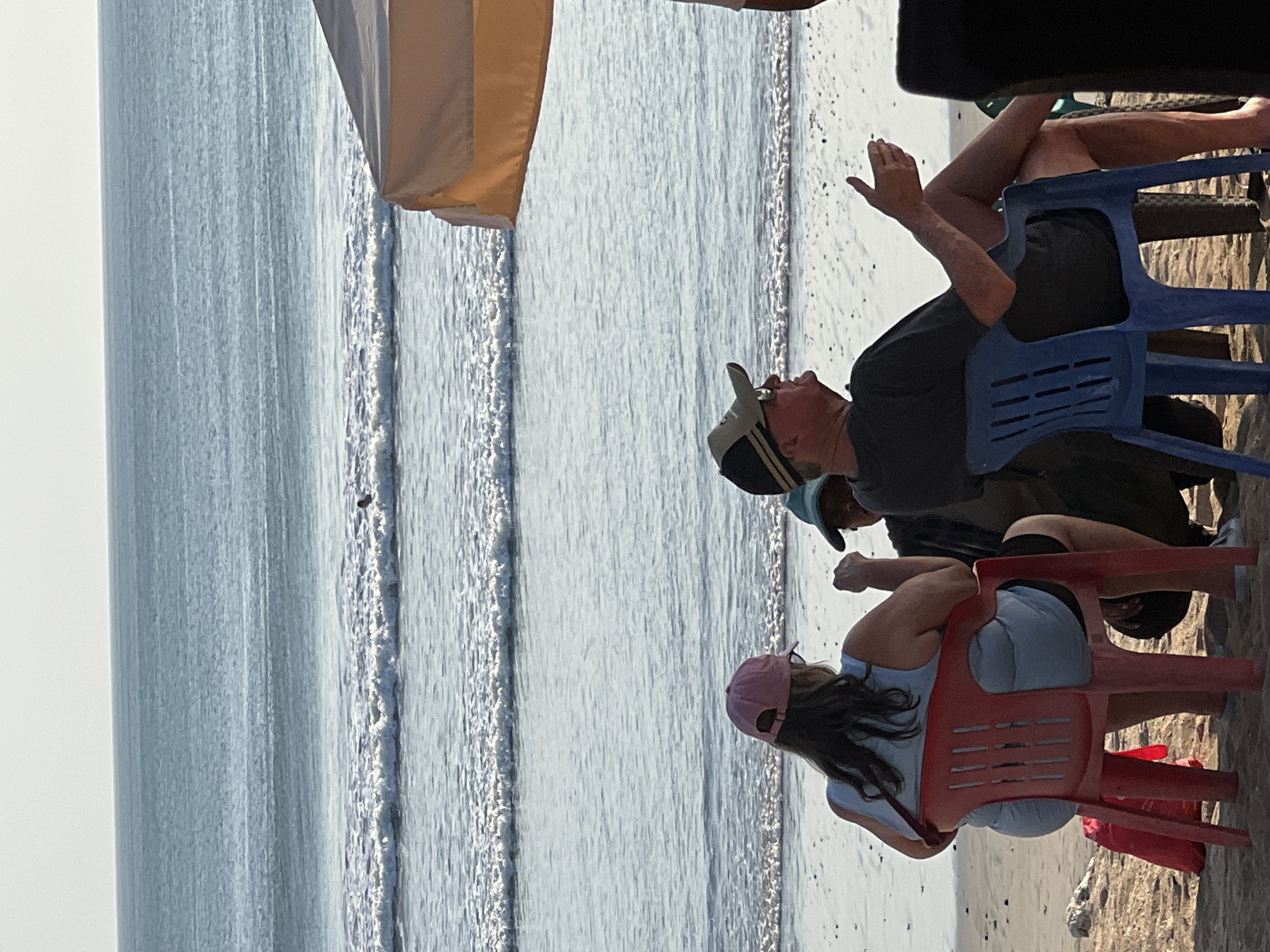 The image depicts a beach scene in Kuta, Indonesia. Three individuals are seated on chairs facing the ocean. In the foreground, a person wearing a pink cap and a light blue tank top sits in a red plastic chair. To their right, a person in a black t-shirt and a baseball cap is seated in a blue plastic chair, gesturing with their hands. Partially visible between them is another individual wearing a teal hat. The ocean stretches out in the background with gentle waves breaking on the shore. Part of a tan-colored awning is visible on the right side of the frame. The ground is sandy beach.