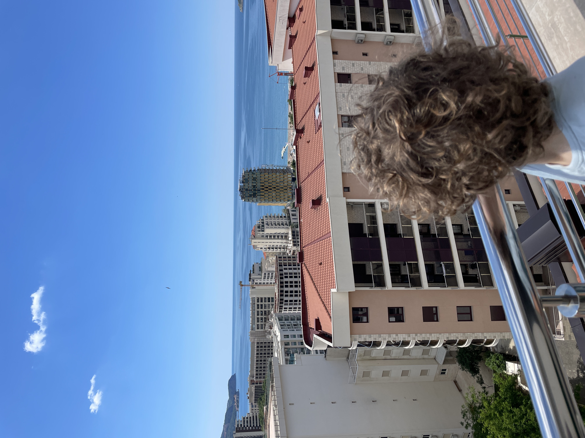 The image depicts a cityscape with a body of water visible in the distance. The perspective is from a high vantage point, possibly a balcony, with metal railings visible in the foreground. To the right, part of a person's head with curly brown hair is visible. Adjacent to this is a multi-story building with a red-tiled roof and beige facade. The building has multiple windows. Beyond this building, a cluster of high-rise buildings is visible along the coast. The water appears calm, and the sky is a clear blue with a few scattered clouds. The location is in the city of Boreti, Montenegro, identified by the coastal city view and the architectural style.