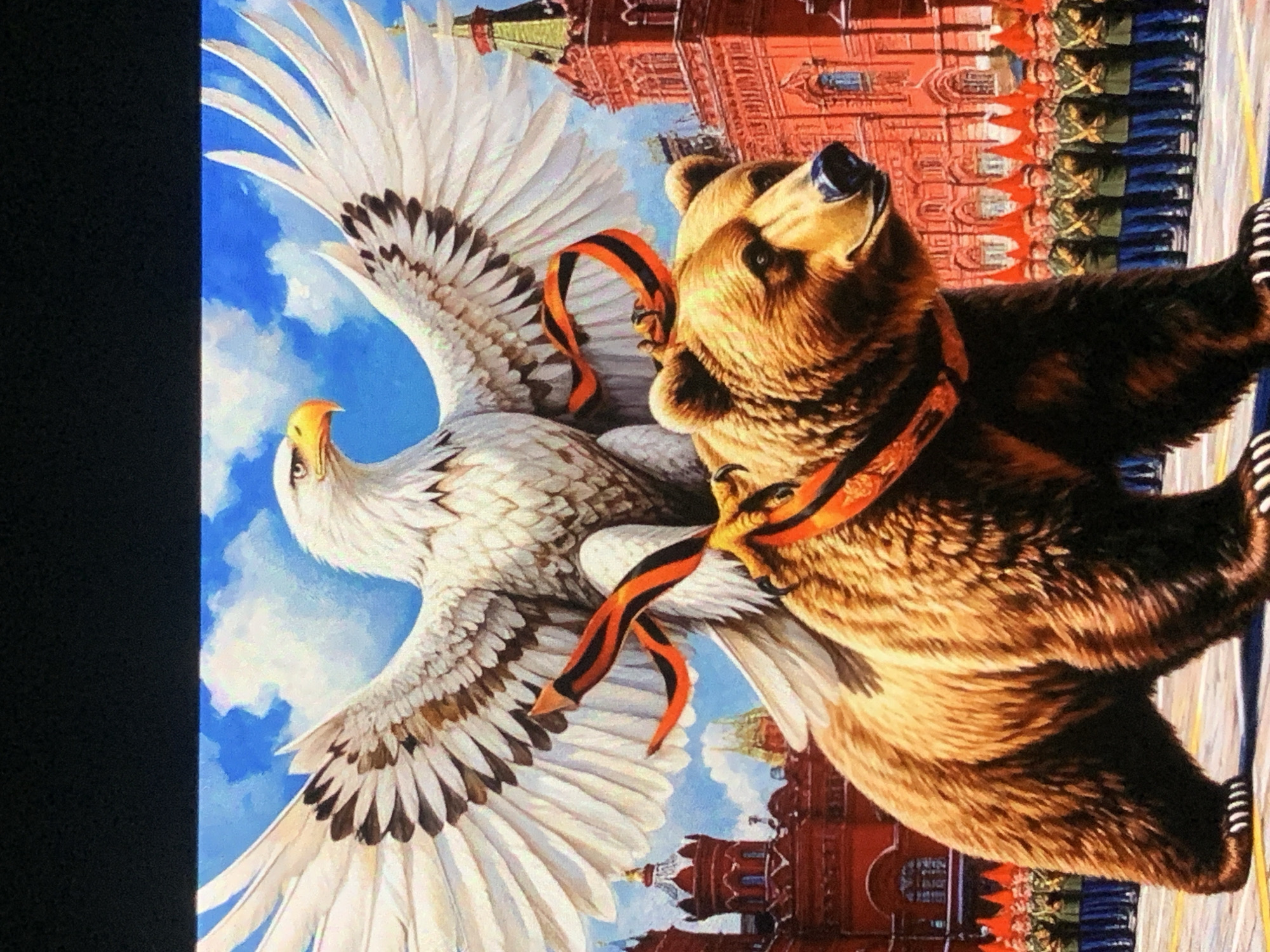 An eagle with outstretched wings perches atop a brown bear. The eagle is white with brown and black markings on its wings. Orange and black ribbons are draped around the eagle and the bear. The bear is looking to the right. In the background, red buildings with towers are visible, as well as a blue sky with clouds. A line of soldiers in green uniforms and red hats are standing at attention in front of the buildings. The scene appears to be a parade on a paved surface. The location is not Sevastopol, Ukraine.