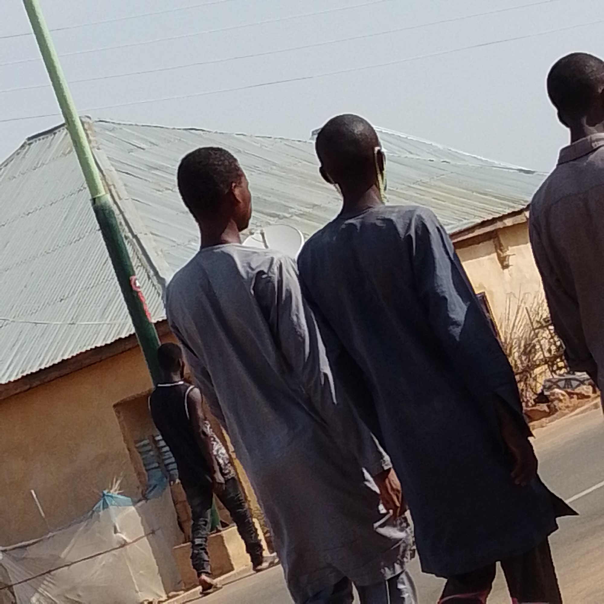 The image shows four individuals walking away from the camera, in what appears to be an outdoor, urban environment in Gombe, Nigeria. The central focus is on three young males, two of whom are dressed in dark blue traditional Islamic attire (buba and sokoto) and one in a grey long-sleeved shirt and trousers. The third young male, to the far left, is wearing a black sleeveless shirt and dark trousers. To the far right, a fourth individual, mostly out of frame, is wearing a light grey shirt and dark trousers.
They are walking on a paved road. In the background, there is a building with a corrugated metal roof and a light brown wall. A tall, thin, green pole with some red marking is visible on the left side of the image. The sky is clear and bright, suggesting a sunny day. There are also some power lines visible in the upper portion of the image. The overall scene depicts a casual moment of people moving through their community.