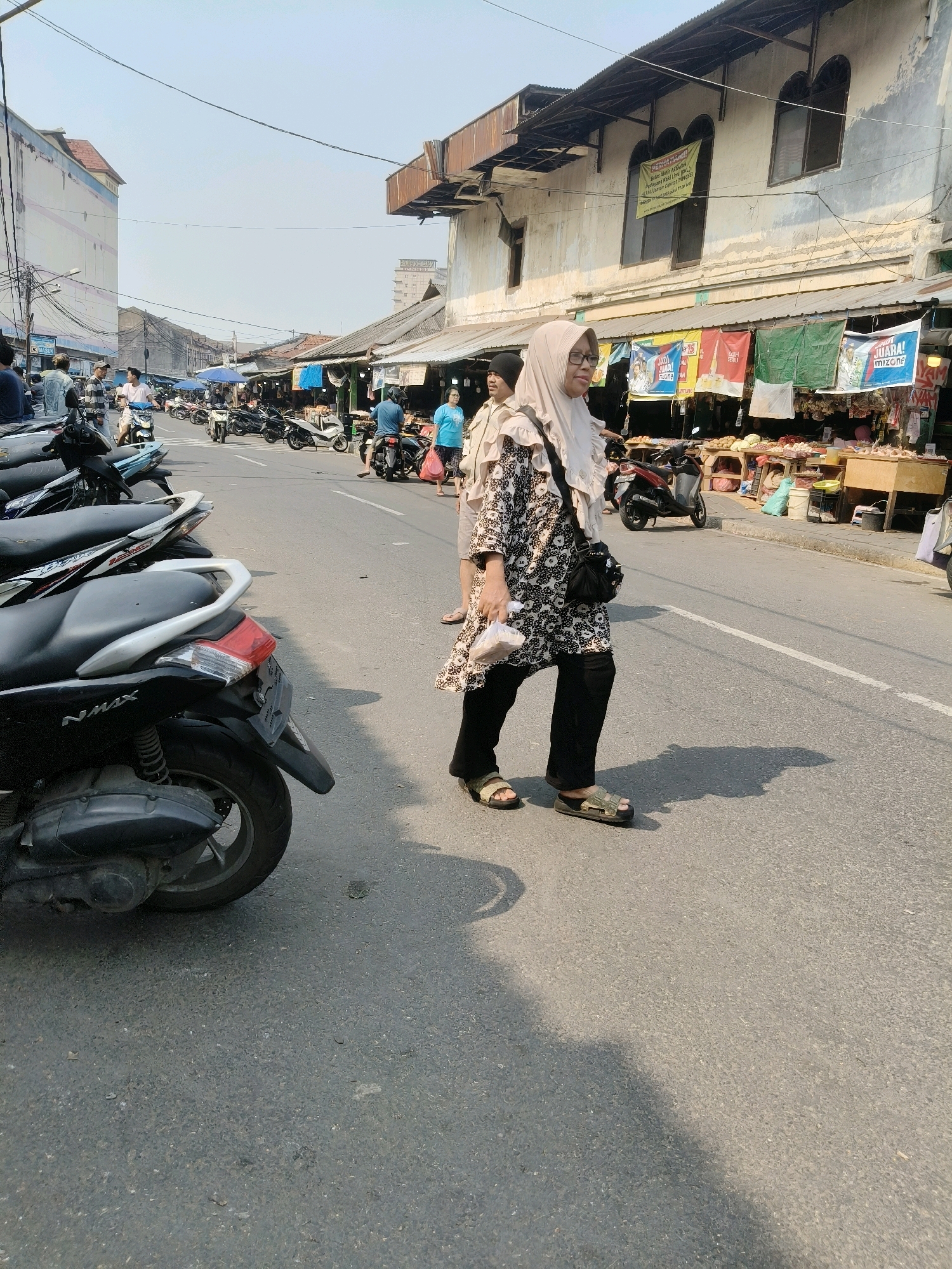The image depicts a street scene in South Tangerang, Indonesia, likely a market area, with a prominent presence of motorcycles and stalls. The foreground features a parked black scooter, identified by the "NMAX" logo on its rear panel. To the right, a woman wearing a light-colored hijab and a patterned tunic walks across the asphalt road, carrying a white plastic bag in her right hand. She is wearing dark pants and sandals. Her shadow stretches across the road, indicating it is daytime.
In the mid-ground, several other motorcycles are parked and some people are seen walking or riding them. Further down the street, market stalls are set up, displaying various goods, and awnings provide shade. A red motorcycle is parked in front of one of the stalls. The buildings lining the street appear to be a mix of older structures with decorative facades and some more modern buildings in the background. The sky is clear and bright. The overall environment suggests a bustling commercial street with everyday activities taking place.
