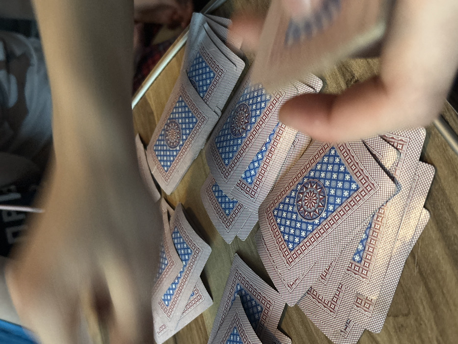 The image depicts an indoor scene in Pervomaiskoe, Russia, focusing on a wooden surface where an activity involving playing cards is underway. Numerous playing cards are scattered across the surface, predominantly in loose piles and individual cards, all displayed with their backs facing upwards. The card backs feature a distinctive blue and red geometric pattern, centered with a star-like motif. Two sets of human hands are visible, both blurred due to motion. In the upper portion of the frame, a blurred forearm and hand are positioned, indicating movement across the scene. In the lower right, fingers from another hand are visible, actively holding or manipulating several cards. The overall impression is one of active engagement in a card game or similar activity.