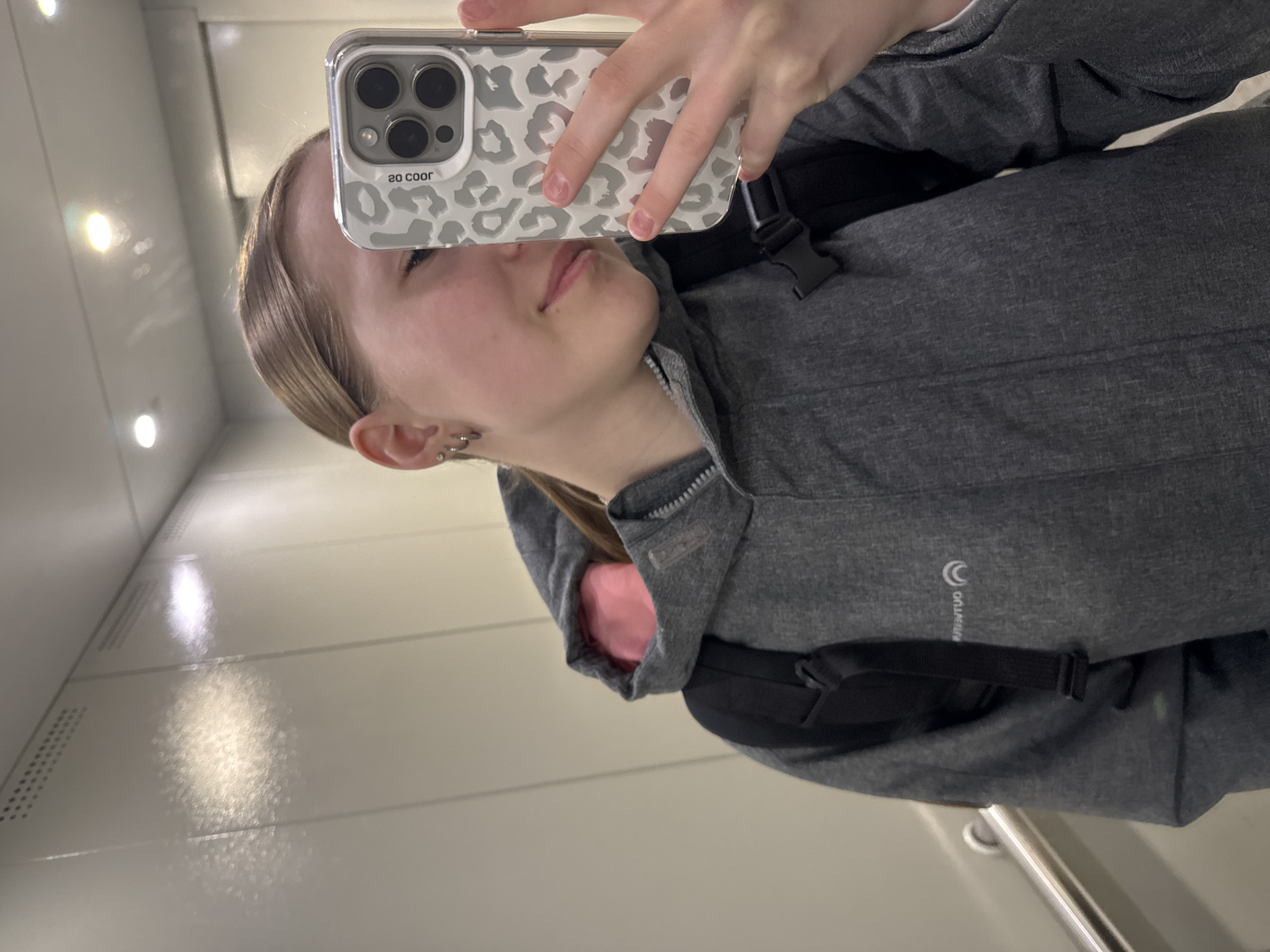 A young individual is captured taking a mirror selfie in an indoor setting in Chaikovsky, Russia. The person, whose gender appears female, is visible from the upper chest upwards, with much of their face obscured by the smartphone they are holding. They have light-colored hair pulled back from their face and display multiple small, silver-toned stud and hoop piercings on their left ear. A neutral to slightly smiling expression is visible on the lower part of their face.
They are dressed in a grey, long-sleeved zippered jacket or hoodie, which features a visible pink lining around the collar and cuffs. A black strap, likely from a backpack or fanny pack, is seen across their upper body. On the right side of the jacket, a brand logo is visible in reflection: a circular 'C' symbol with an inner circle, and the word "CHAMPION" written vertically below it (appearing reversed as "OITNYUD" in the image).
The smartphone used for the selfie is encased in a clear cover adorned with a white and grey leopard print pattern. The phone itself has a prominent camera module with three distinct lenses. Text on the phone case, visible in reflection, reads "2o coor", which is likely "so cool" when viewed correctly.
The background comprises reflective, light-colored wall panels or a mirror, illuminated by bright artificial overhead lights, indicating an indoor environment such as an elevator, hallway, or a modern room.