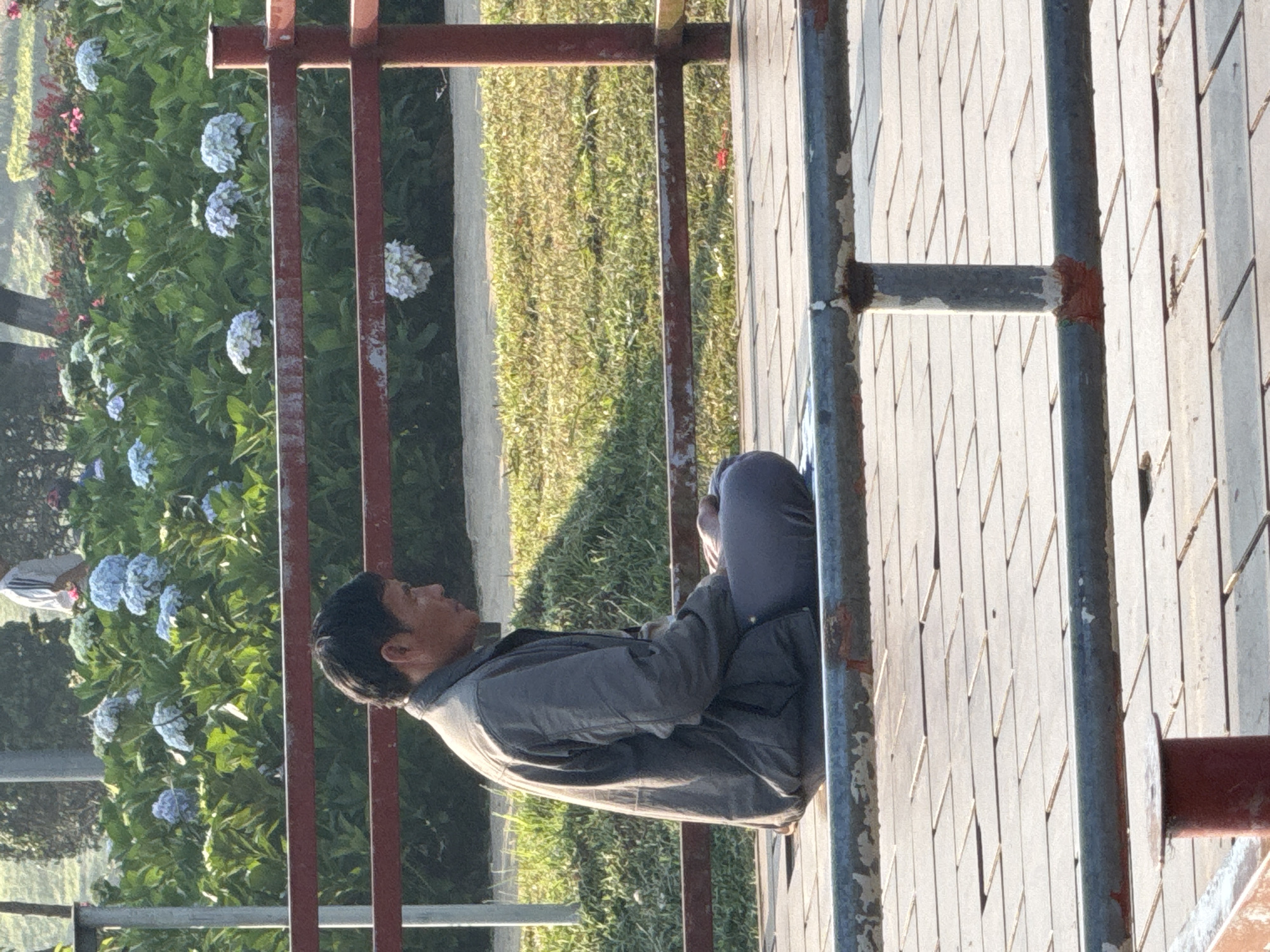 An Asian man sits cross-legged on a concrete tile bench in Da Lat, Vietnam. He wears a gray jacket and pants. The bench is surrounded by a rusty metal railing. Behind the bench, there is a flower bed with blue hydrangeas and green grass.