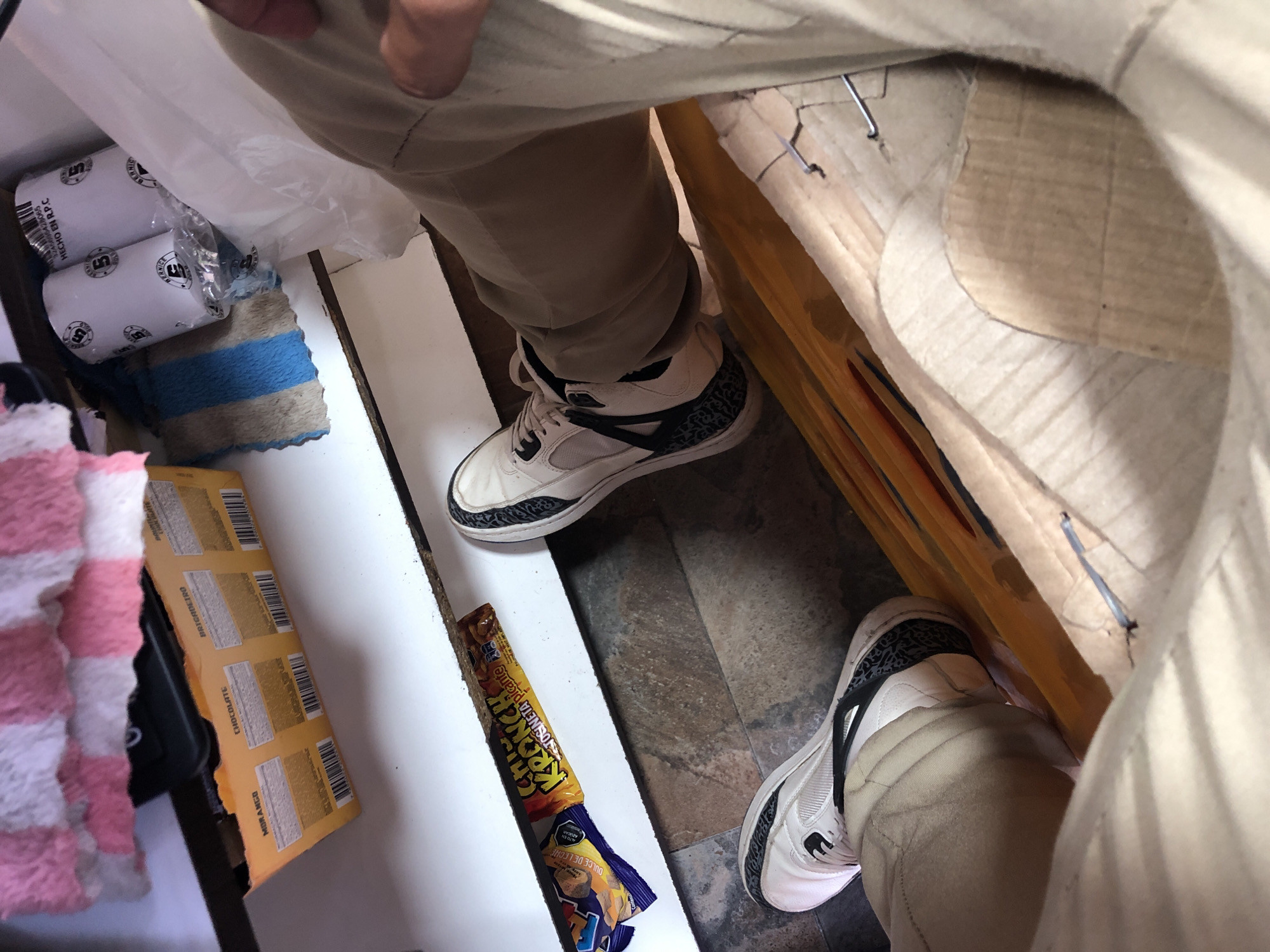 The image shows a person standing in what appears to be a room with a tiled floor. The person is wearing beige pants and white sneakers with black accents. In the background, there are various items including rolls of what may be medical tape, a blue and grey striped cloth, a box of what appear to be flavored wafers, and a candy bar wrapper. The environment appears cluttered with items placed around the person's feet. The location within the city of Juan Griego, Venezuela, is not discernible from the provided image.