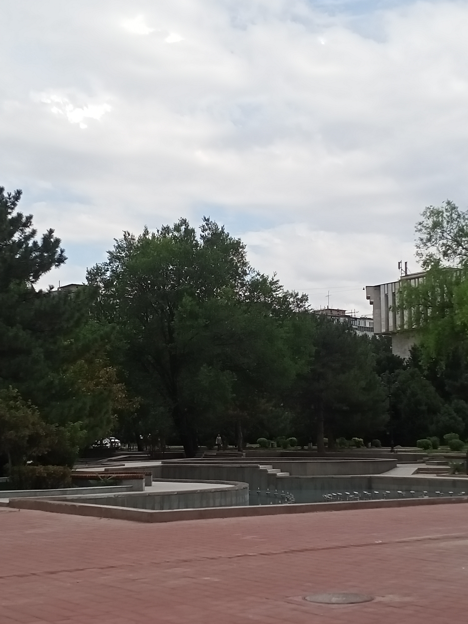 A medium shot captures a fountain in a city park with buildings in the background. The fountain, which appears to be dry, is constructed with multiple tiered levels and concrete borders. The surrounding area is paved with reddish bricks. To the left and right of the fountain, lush green trees stand tall, with some buildings partially visible behind them. The sky above is overcast with a few patches of light. A person can be seen in the distance, walking through the park. The overall scene depicts a calm, public outdoor space in Bishkek City, Kyrgyzstan.